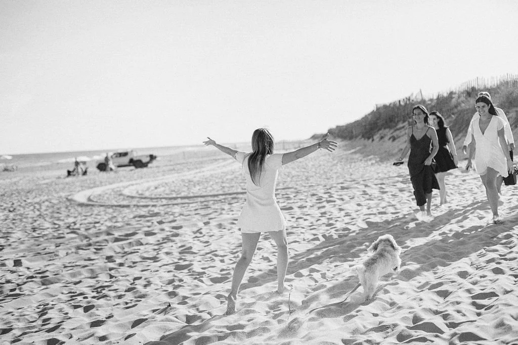 A woman standing on a beach with her back to the camera, arms outstretched, facing a group of three women walking towards her, and a dog running alongside them, with a few boats in the distance on the sand and a dune line in the background.