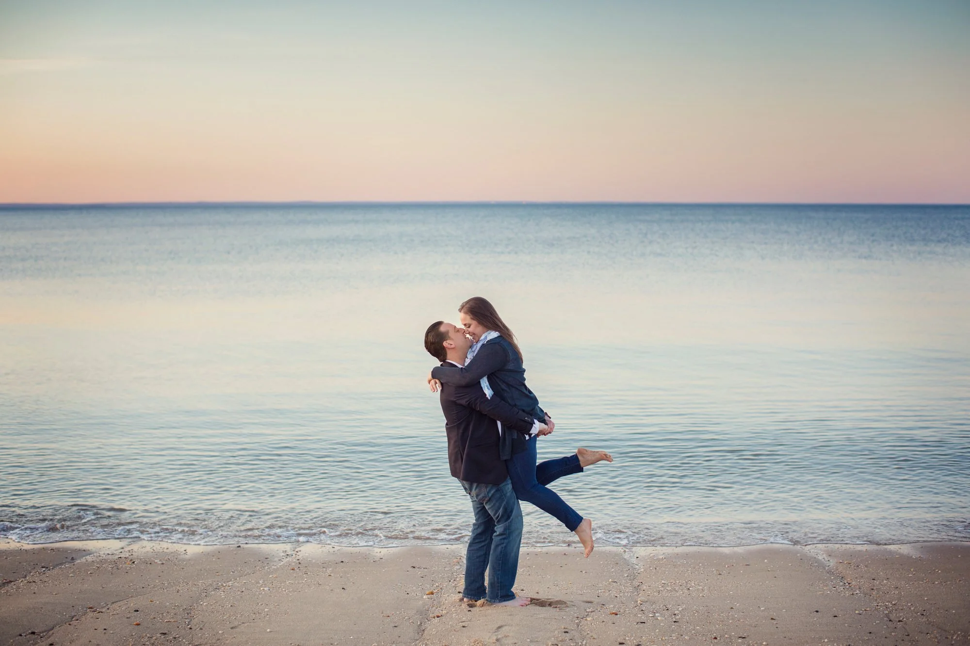 A couple on the beach with the man lifting the woman in his arms, both smiling with the ocean and a pastel-colored sky in the background.