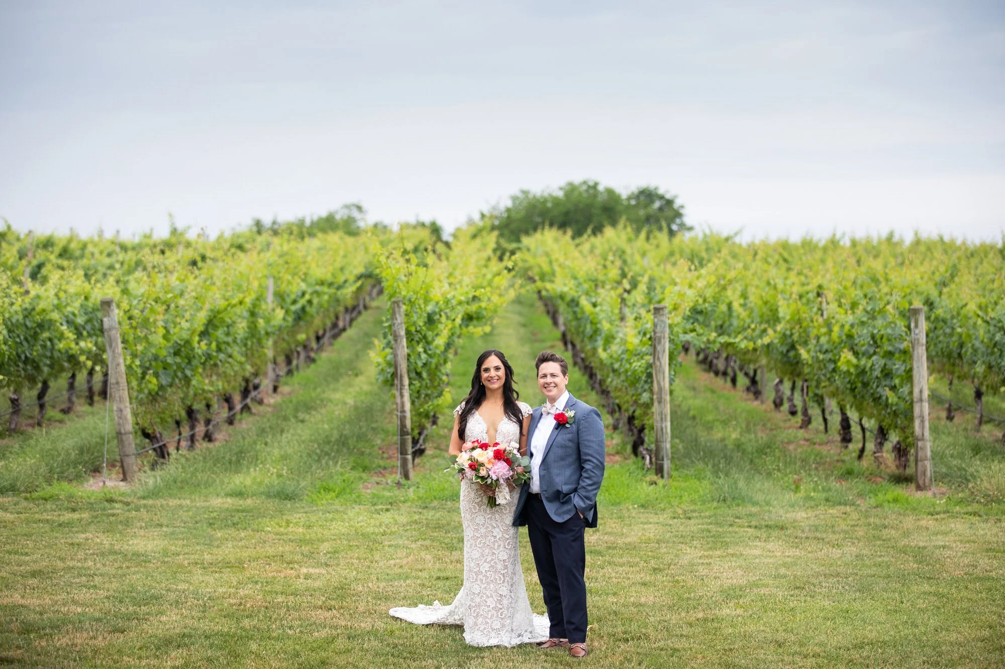 A bride and groom standing in a vineyard, smiling, with the bride holding a colorful bouquet, both dressed in wedding attire.