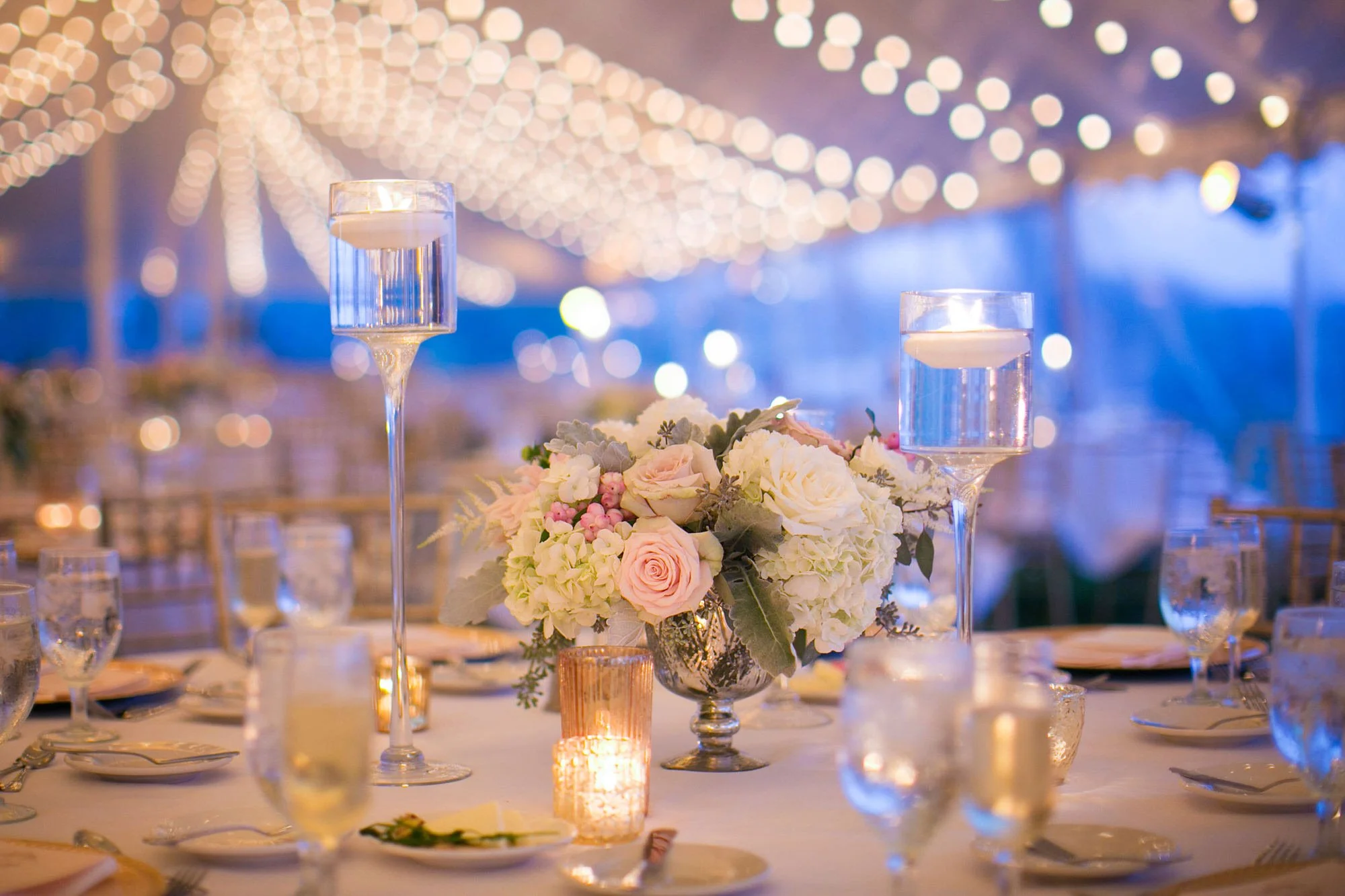 Elegant wedding reception table with floral centerpiece, candle, and floating candles in tall glass holders, under string lights in a festive outdoor setting at dusk.