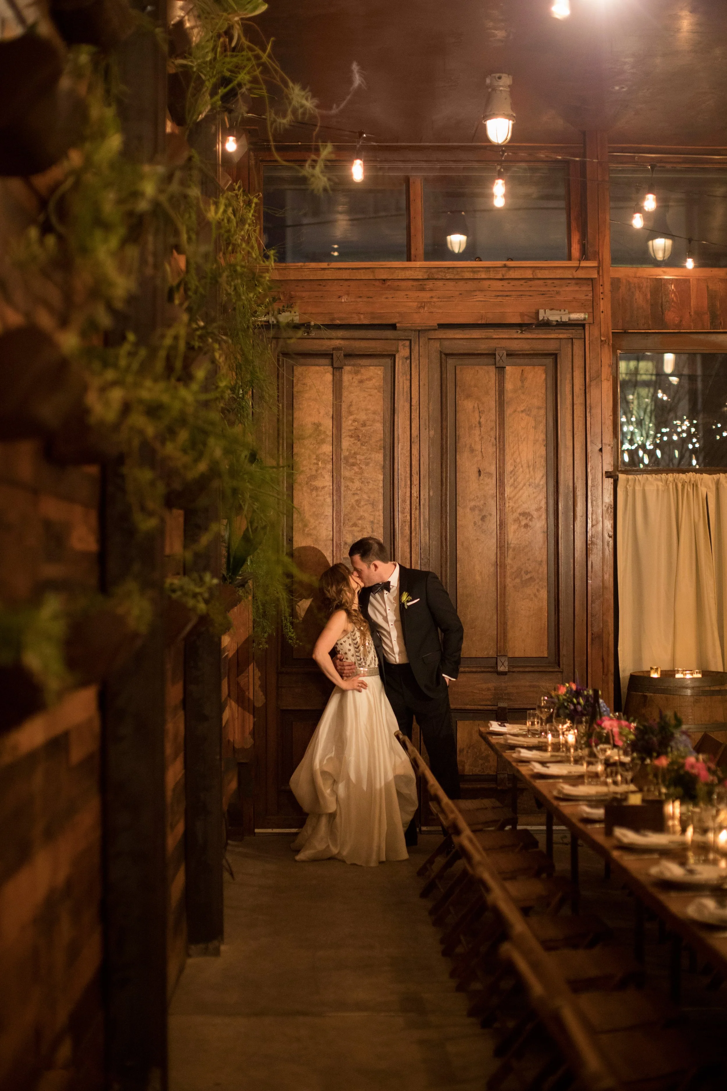 A bride and groom sharing a kiss at their wedding reception in a warmly lit, rustic wooden room with a long table decorated with flowers and candles.