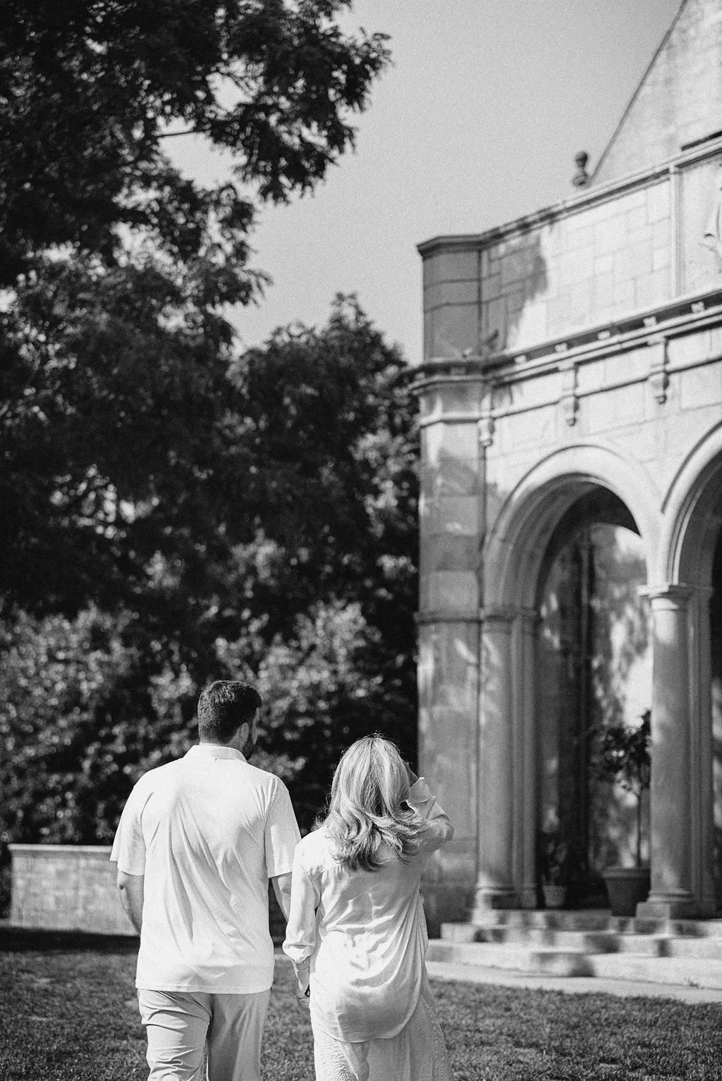 A black and white photo of a man and woman walking together outside near a large stone building with columns, surrounded by trees.