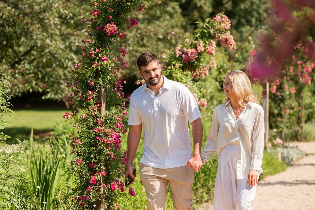 A man and a woman walking hand-in-hand on a garden path with flowering bushes and trees around them, smiling and enjoying each other's company.