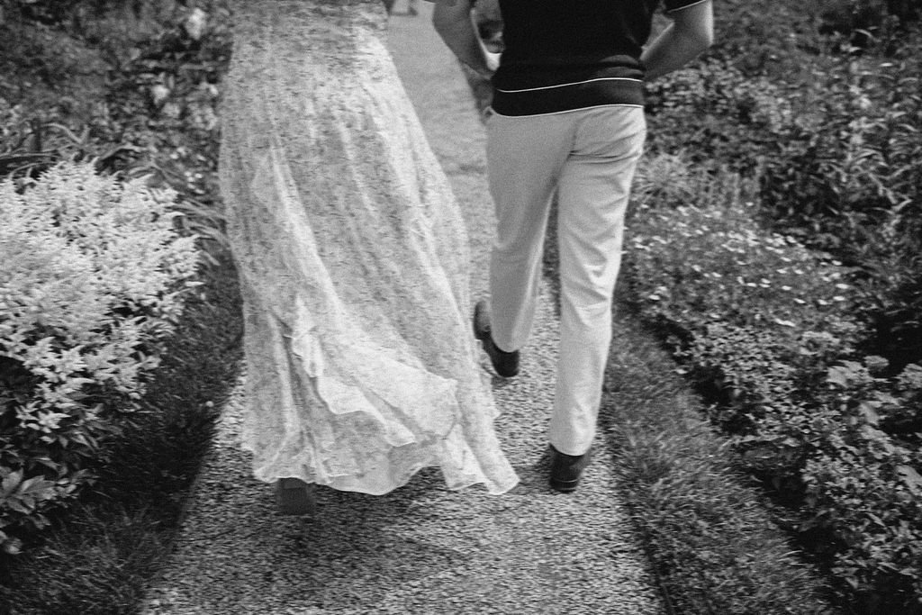 A man and a woman walking along a garden path, viewed from behind. The woman is wearing a long dress and the man is dressed in light-colored pants and a dark shirt.