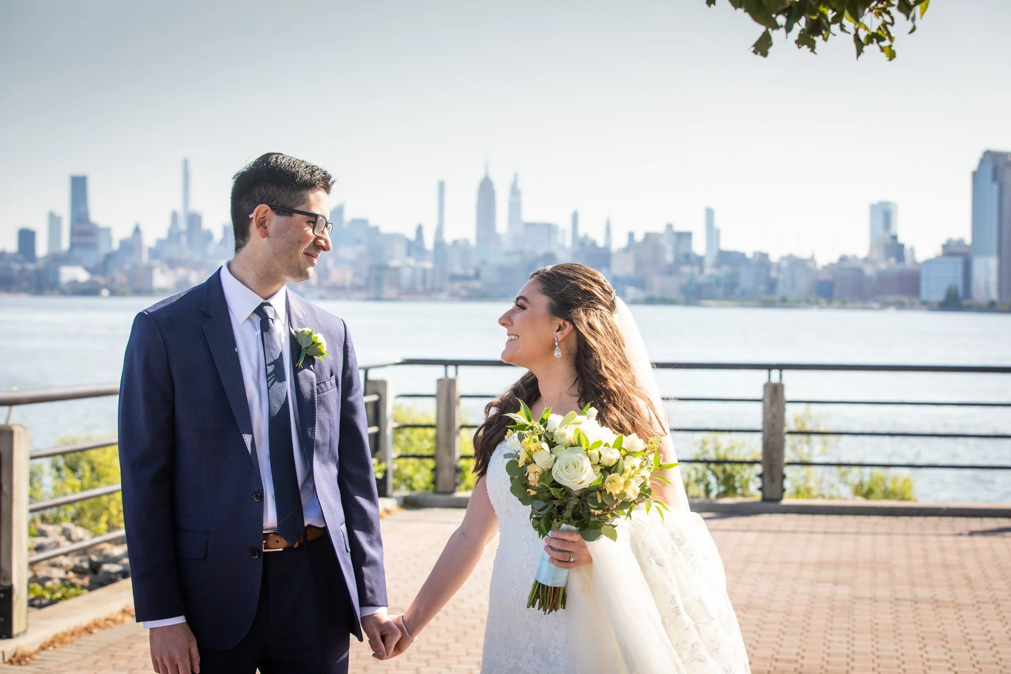 A bride and groom holding hands, standing outdoors by a body of water with a city skyline in the background, during a wedding ceremony.