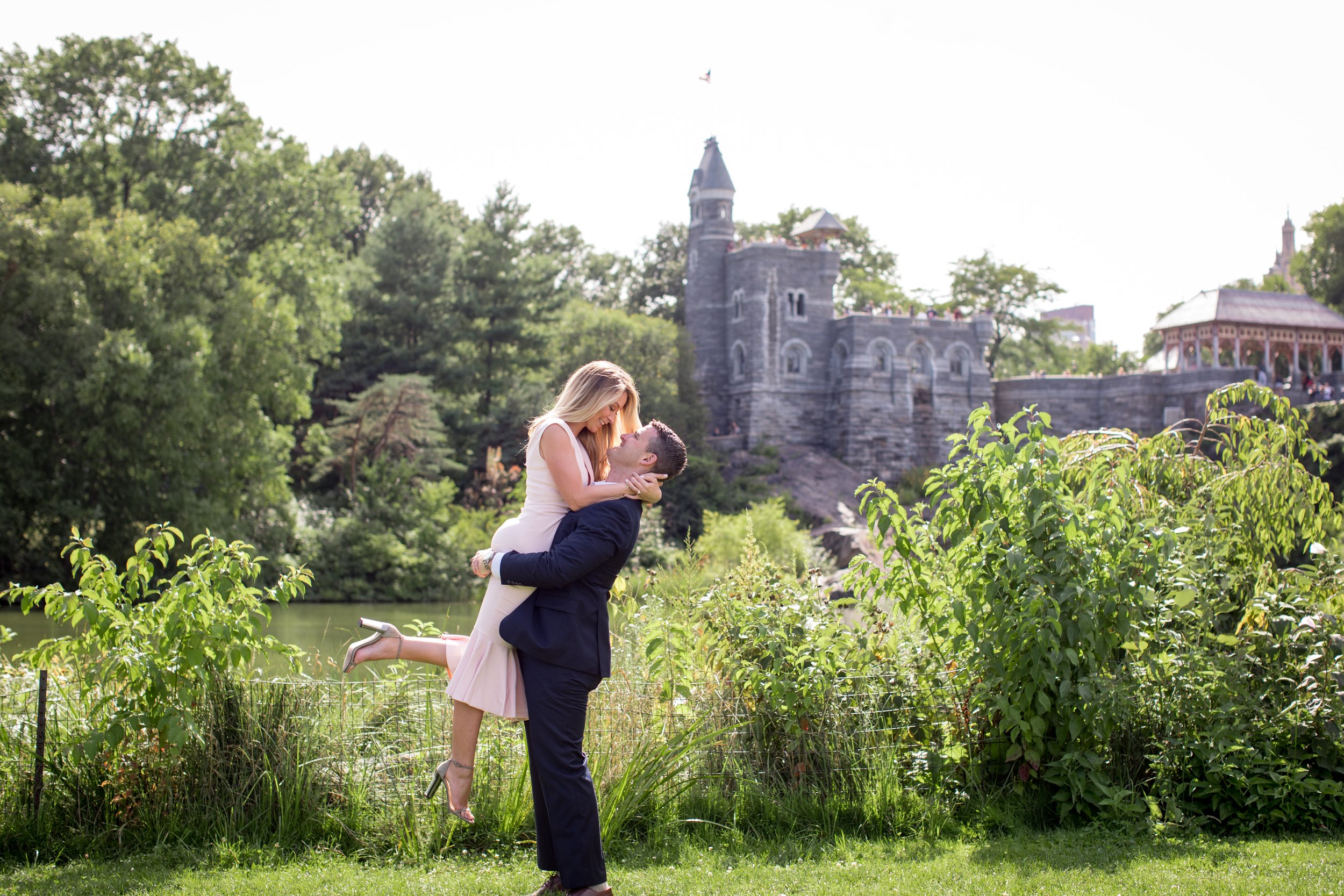 A couple in formal attire sharing a romantic moment in a park with a stone castle-like building in the background.