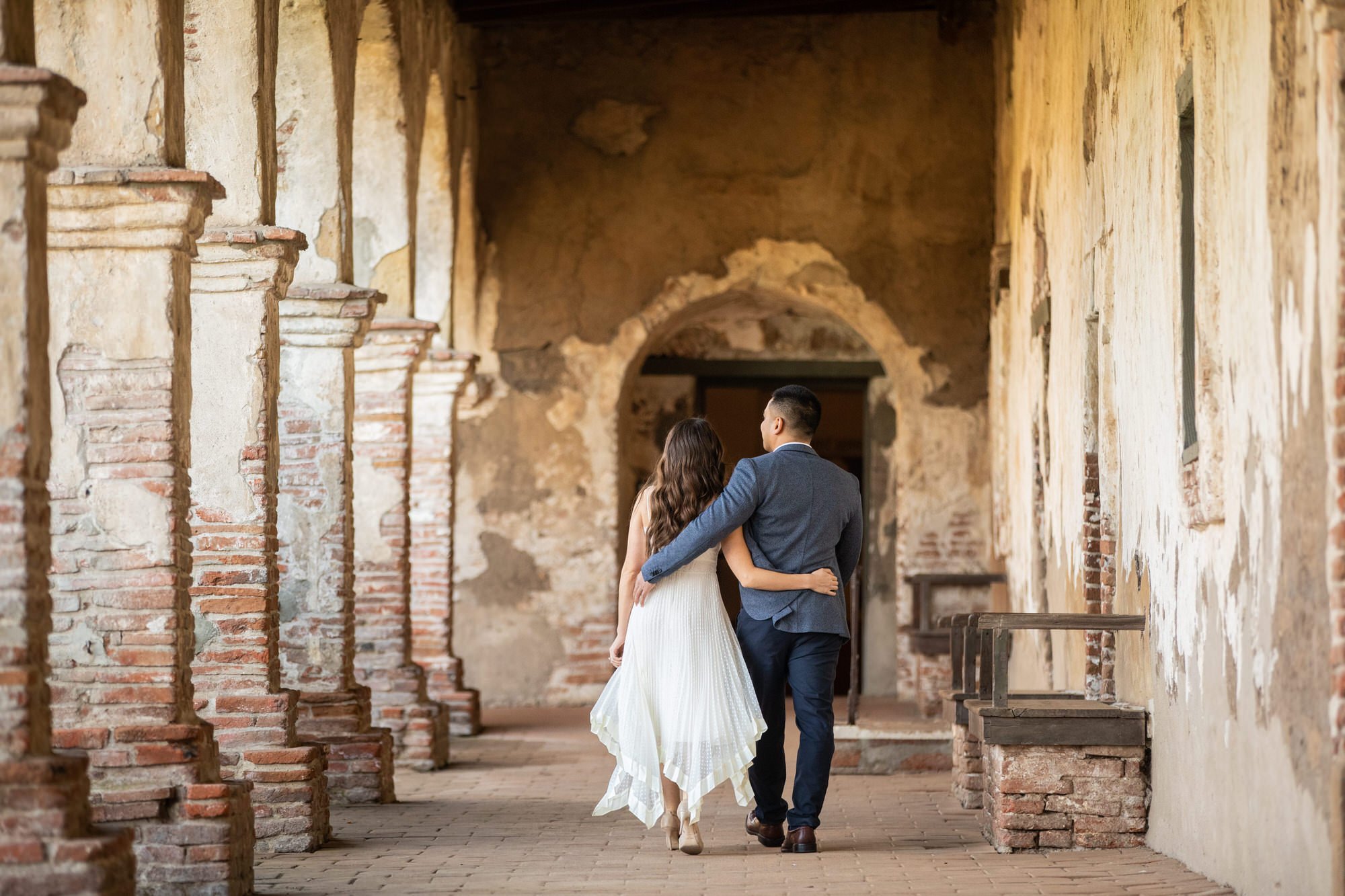 A couple walking arm in arm through a rustic, historic hallway with weathered brick and plaster walls, and arched doorways.