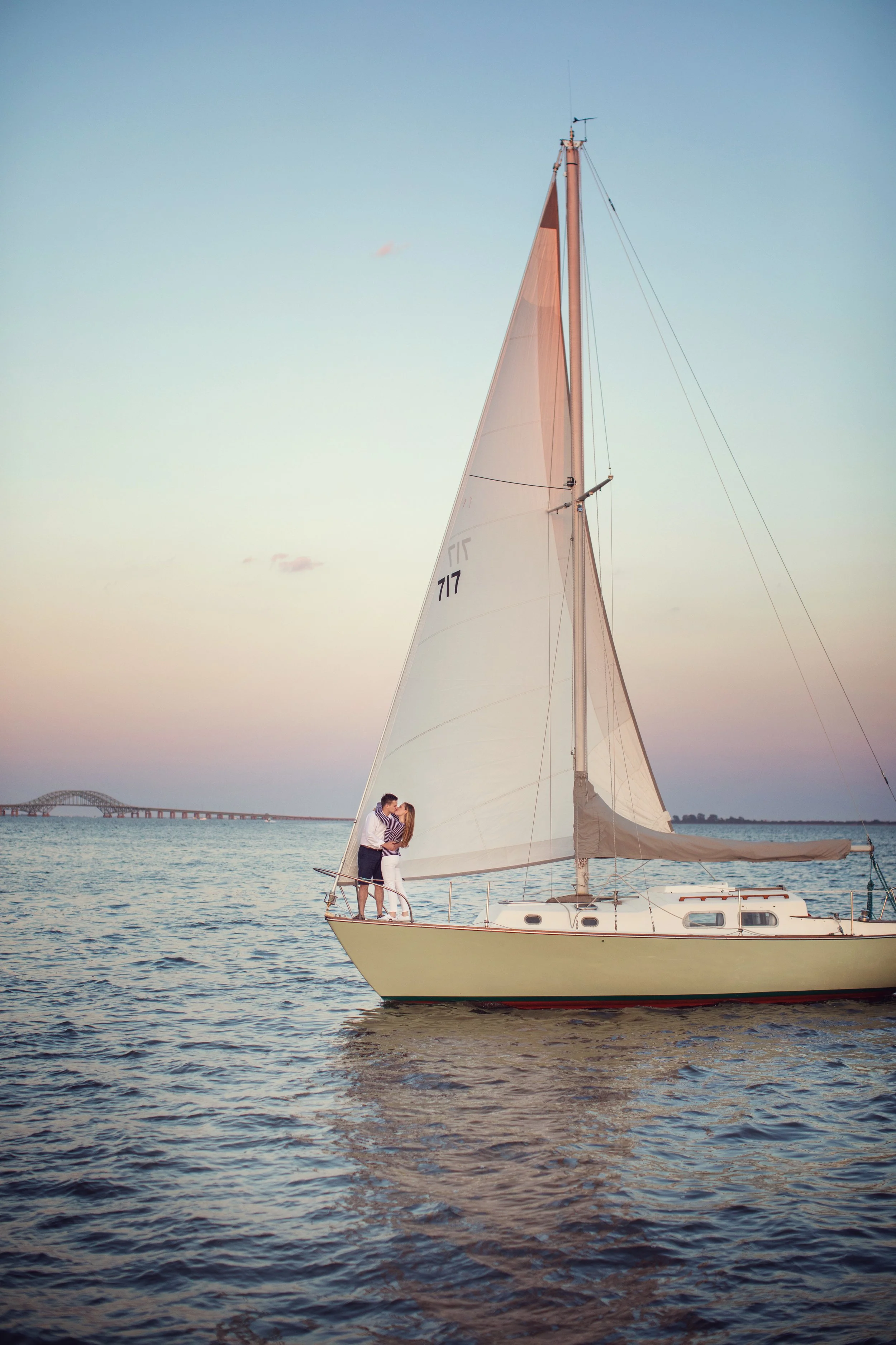 A couple kissing on the bow of a sailboat on calm water at sunset with a bridge in the background.