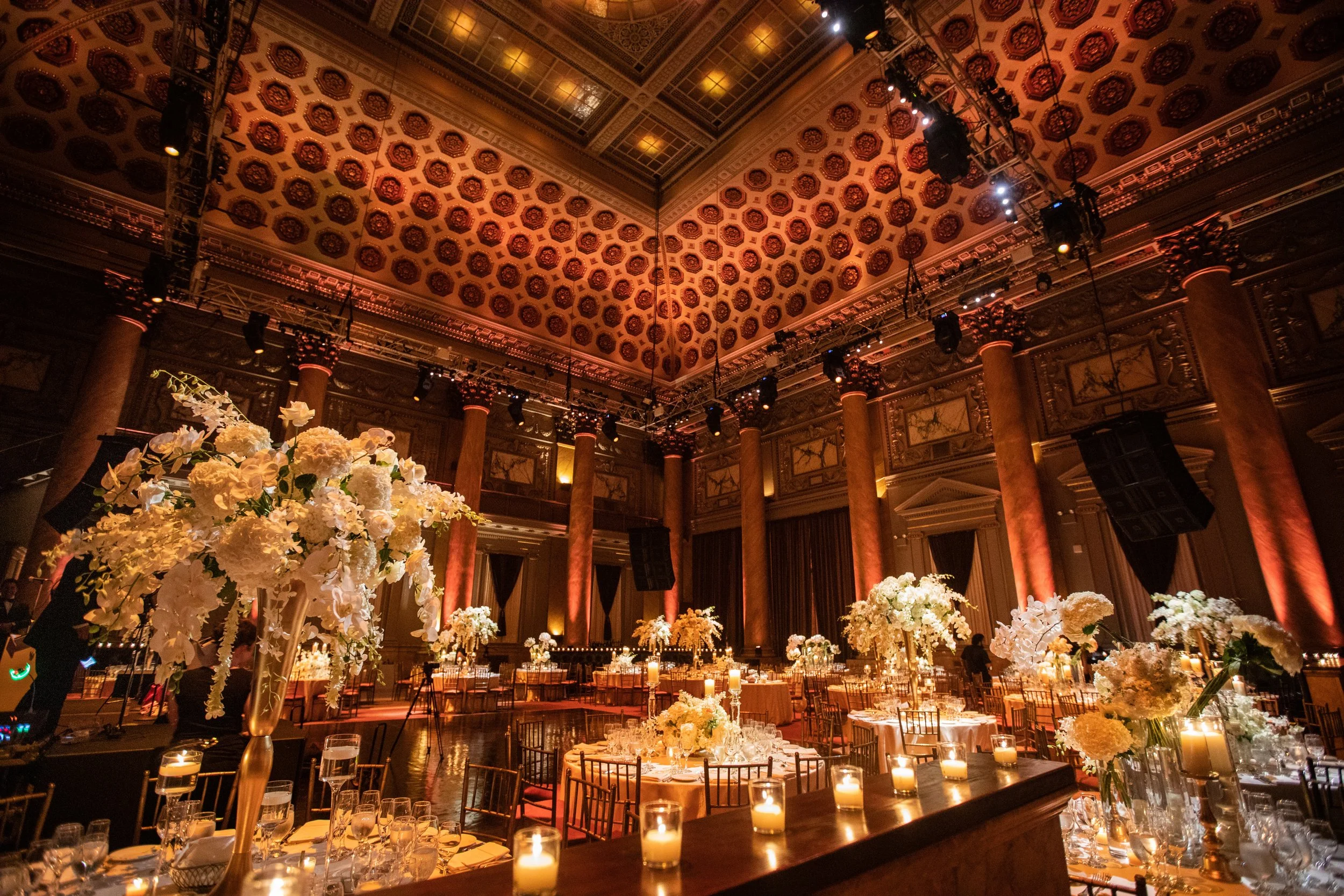 Elegant banquet hall decorated with tall floral arrangements of white flowers on tables, candles, and warm lighting, with ornate ceiling and columns.