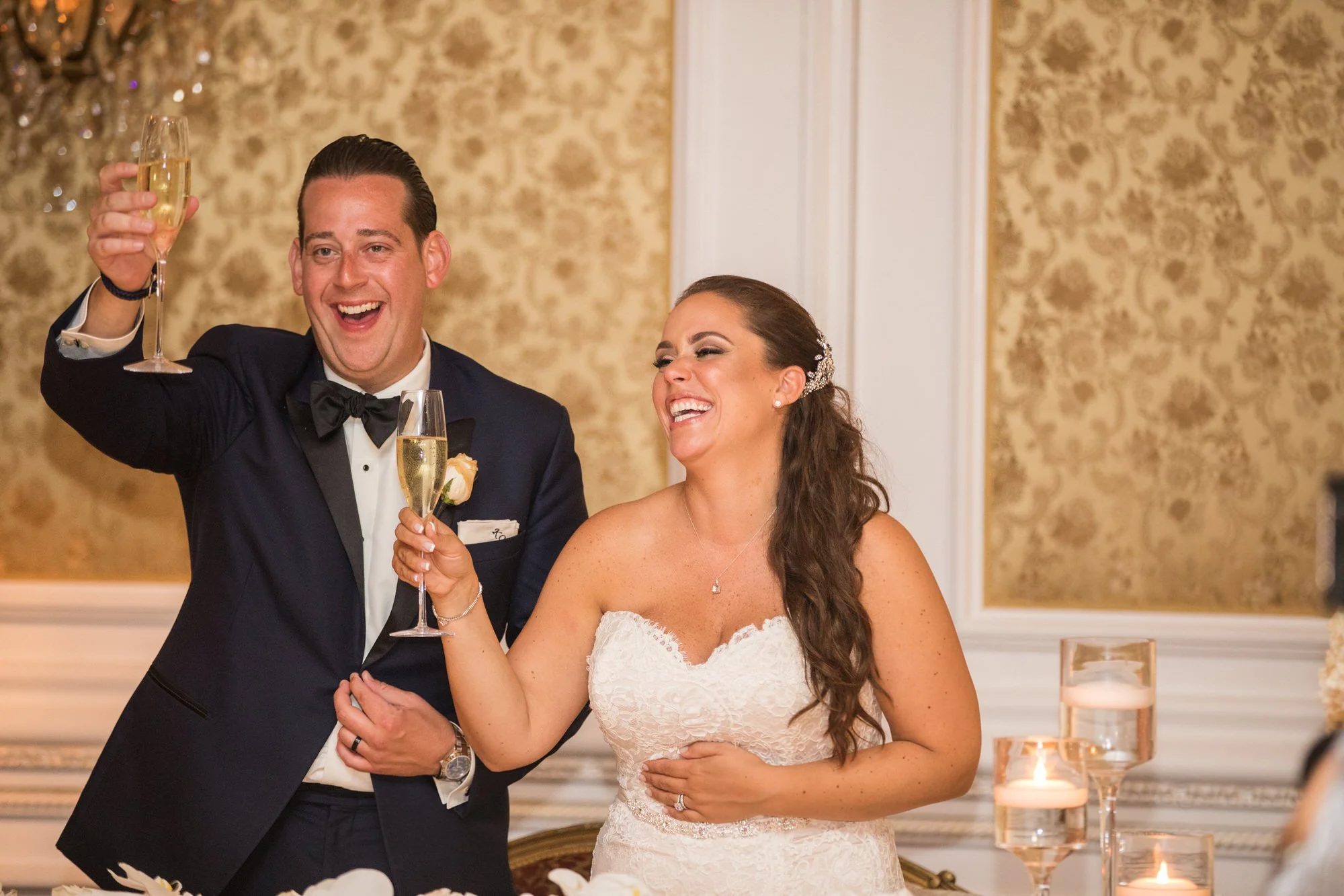A newlywed couple dressed in wedding attire laughing and raising champagne glasses during a celebration.