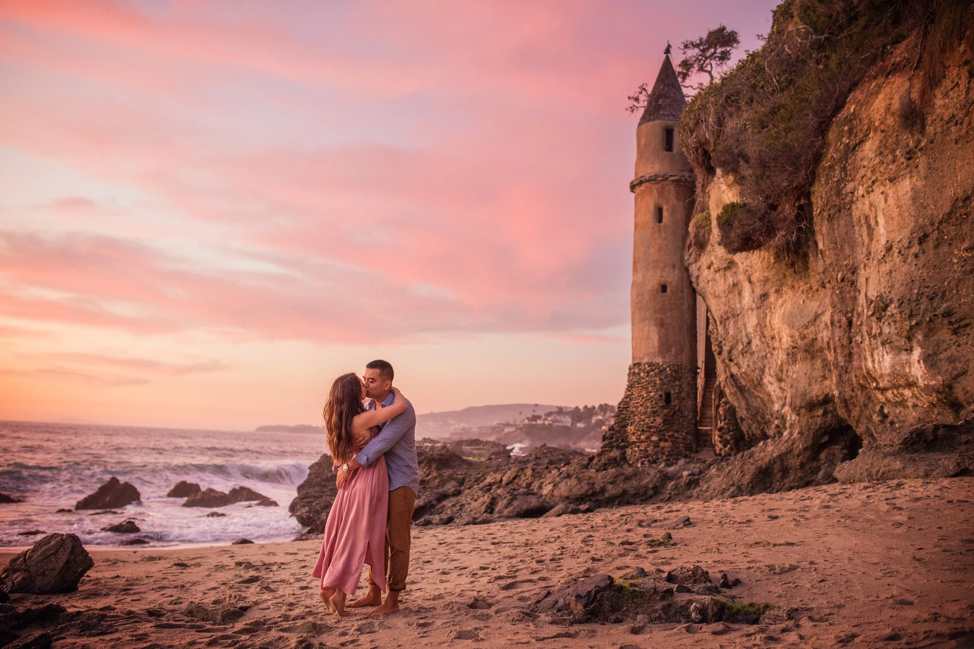 A couple embracing on a beach at sunset with a castle tower on a rocky cliff in the background.
