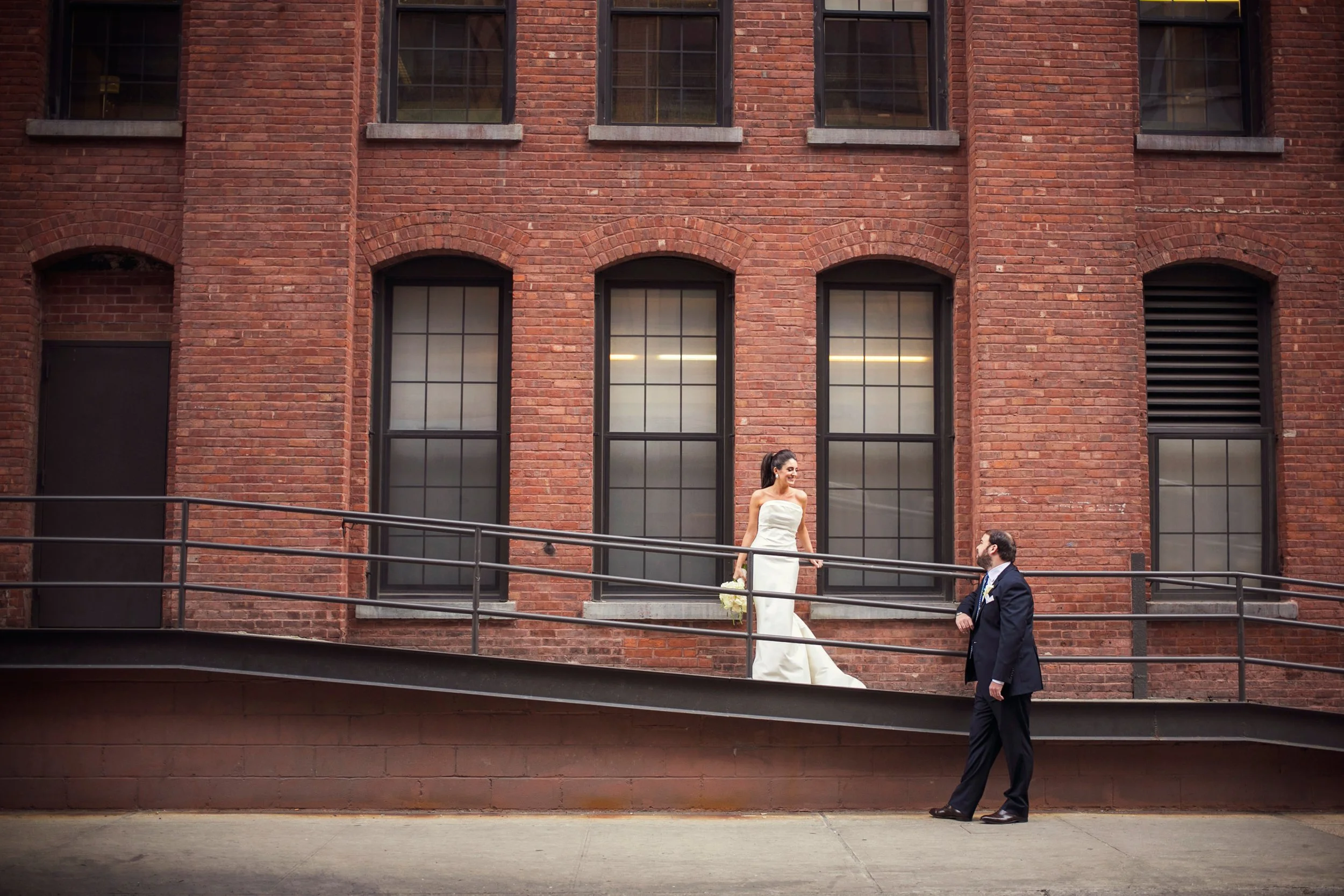 A newlywed couple stands on an urban brick building's ramp; the bride in a white wedding dress holding a bouquet, smiling at the groom who is dressed in a dark suit. The background shows large, black-framed windows and a brick wall.