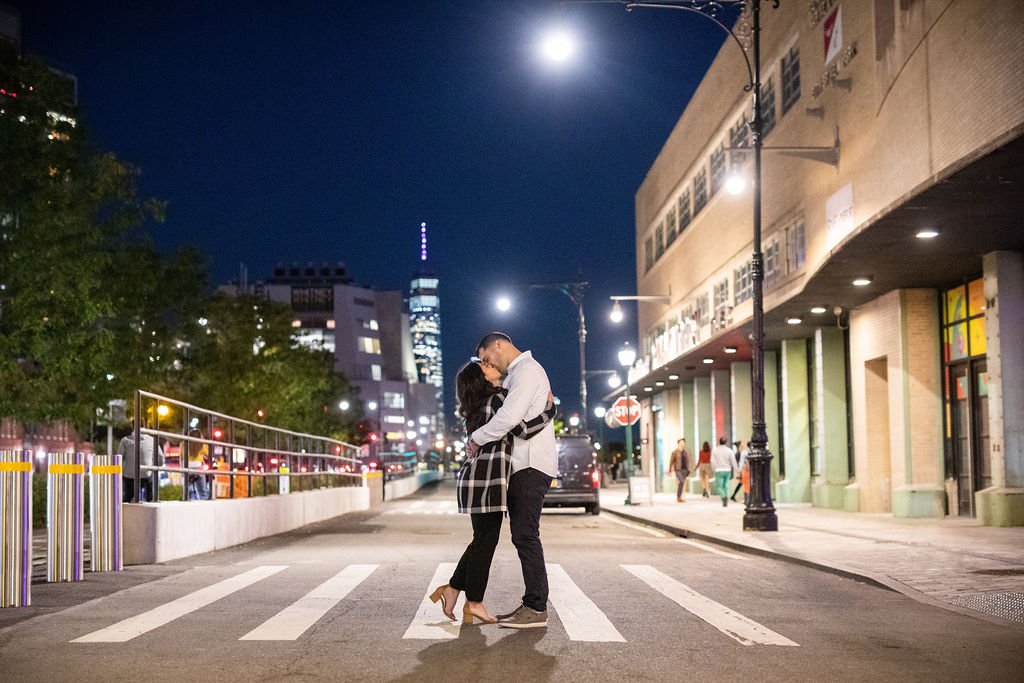 A couple embracing on a city street at night, standing on a crosswalk, with buildings and streetlights illuminated in the background.