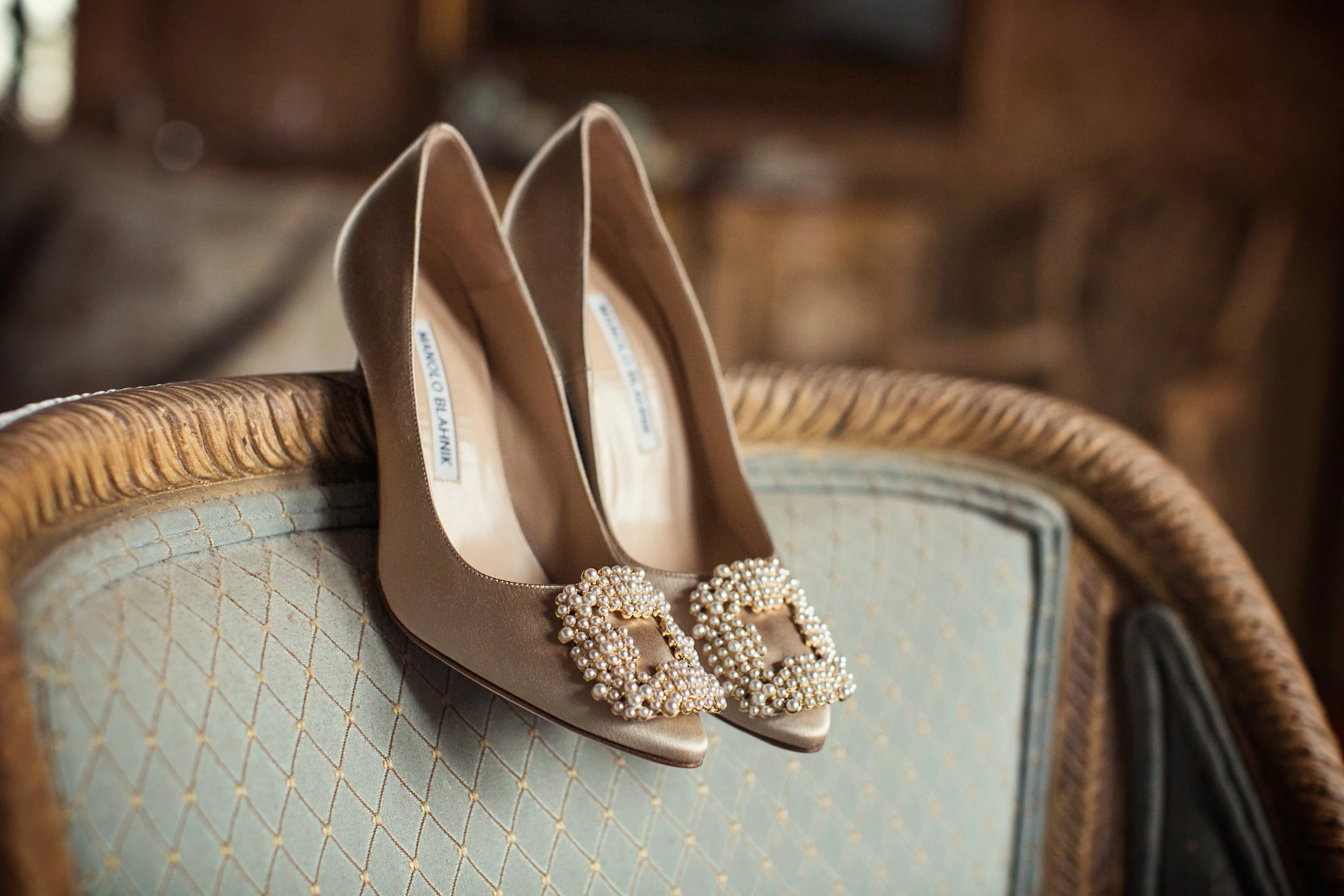 A pair of beige high-heeled shoes with pearl embellishments on the front, resting on a vintage upholstered chair with a wooden frame.