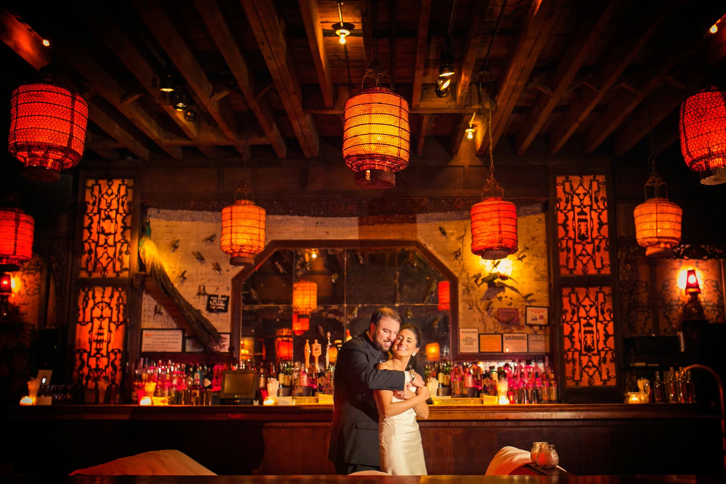 A happy couple hugging in a warmly lit bar with red lanterns hanging from the ceiling and a large mirror behind the bar reflecting the scene.