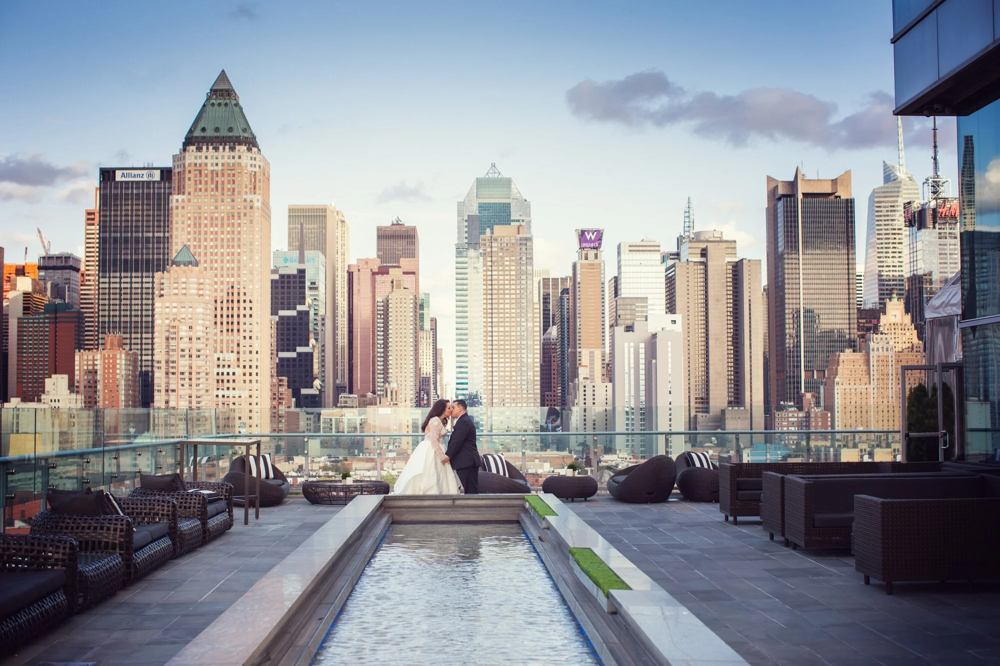 A bride and groom holding hands and about to kiss on a rooftop balcony with a city skyline of tall skyscrapers in the background. The rooftop features outdoor seating, a small pool, and a glass railing.
