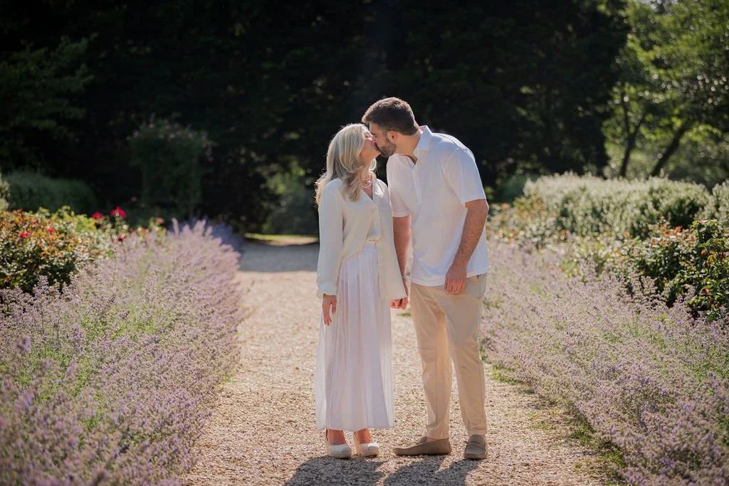 A couple sharing a kiss on a gravel garden path surrounded by flowering bushes and trees.