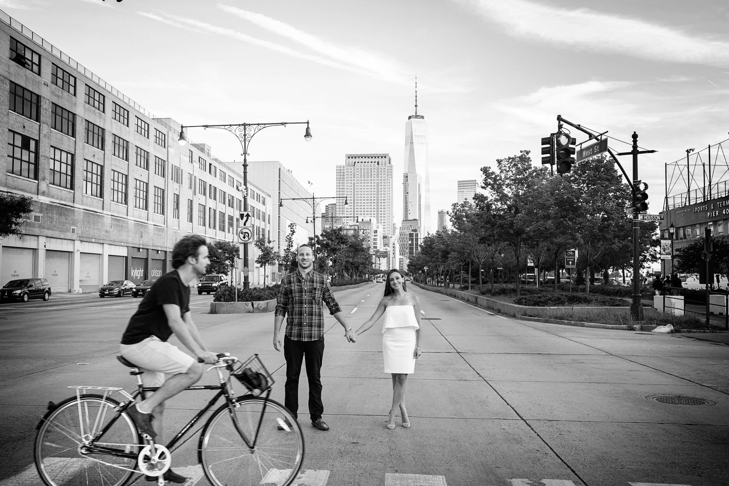 A man and woman holding hands and walking across the street with a cyclist passing by in an urban cityscape with tall buildings and trees, black and white photo.