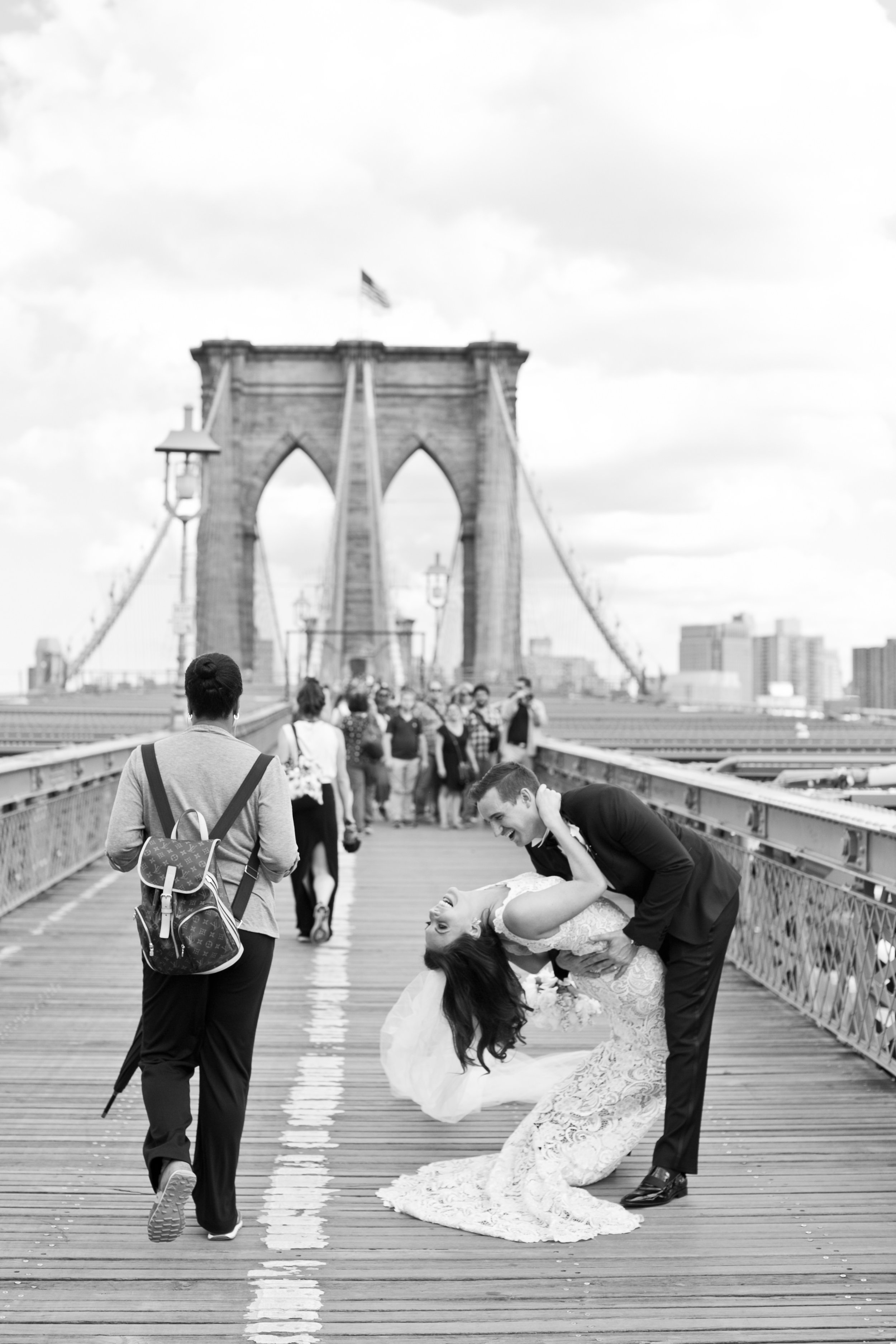 A couple in wedding attire dancing on the Brooklyn Bridge while pedestrians walk in the background, with the bridge's towers visible.