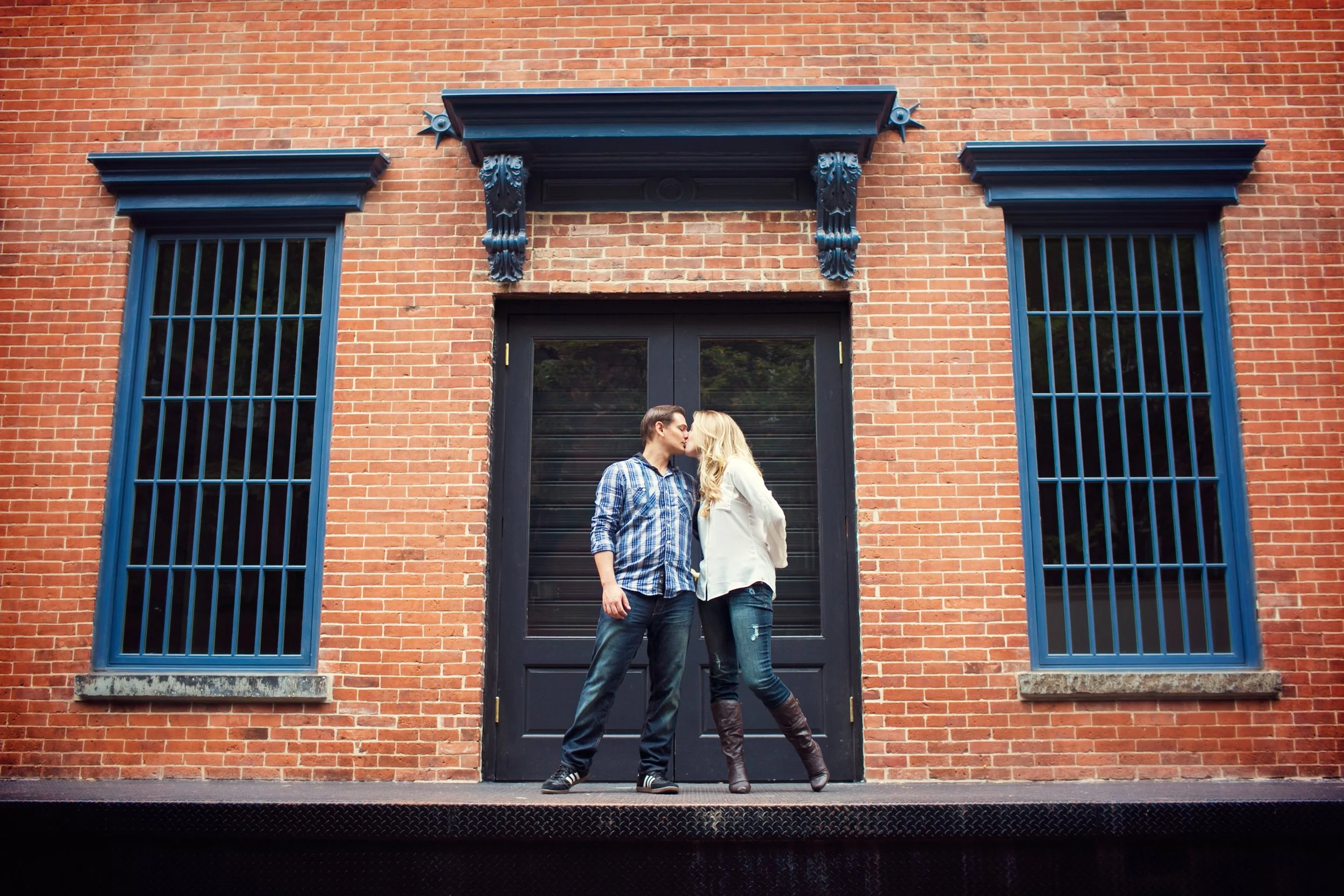 A couple sharing a kiss in front of a brick building with blue window frames and black door.