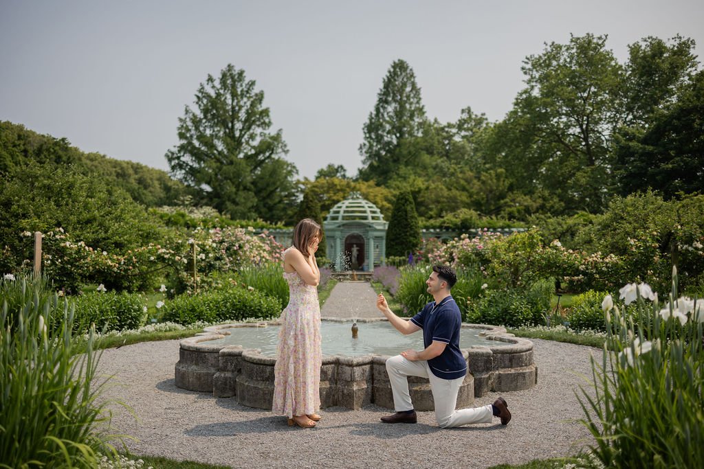 A man proposing to a woman in a garden with a fountain, surrounded by flowers and trees, with a gazebo in the background.