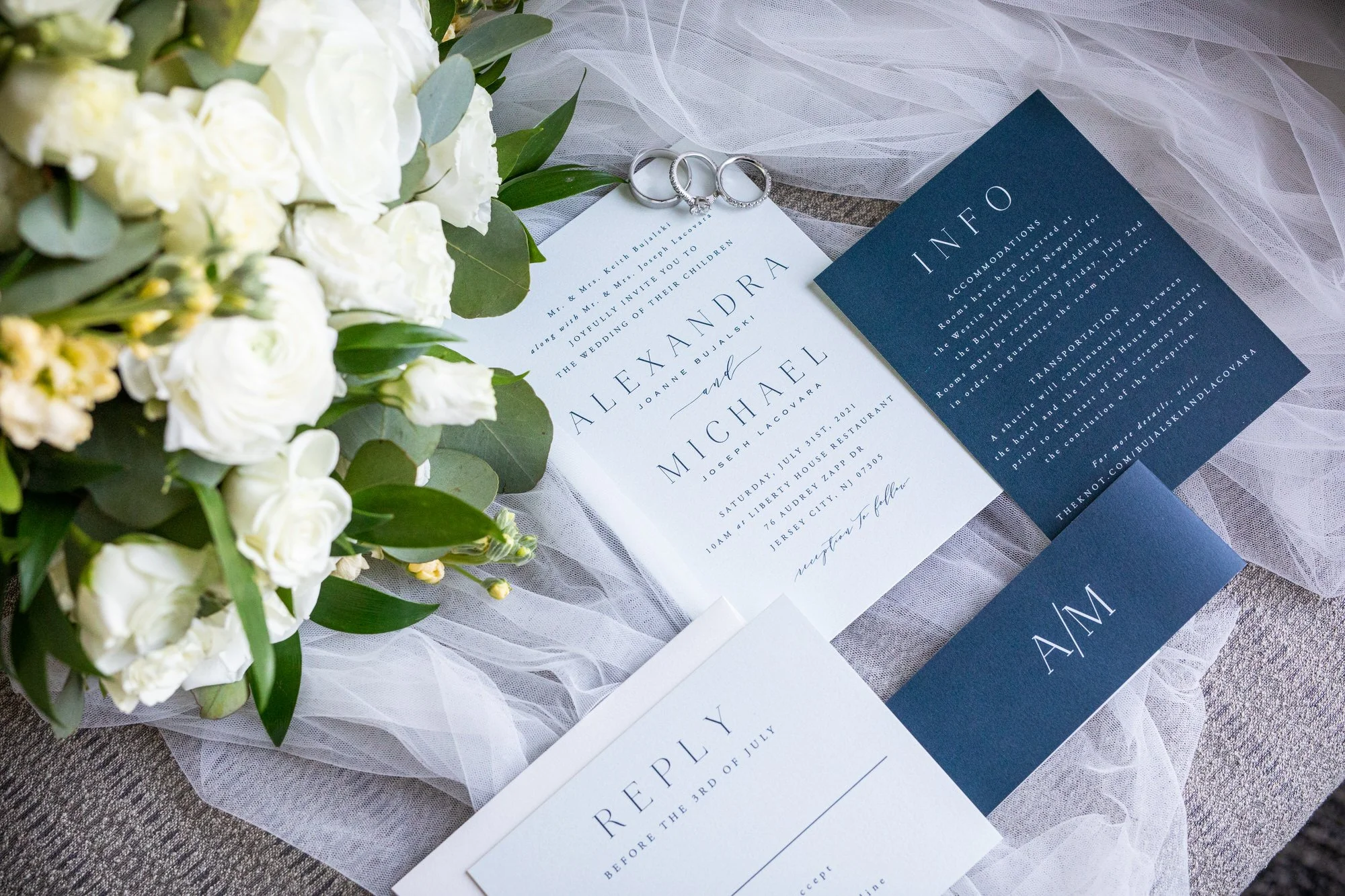 Wedding invitation suite with rings, floral bouquet, and an accommodation card on a sheer fabric surface.