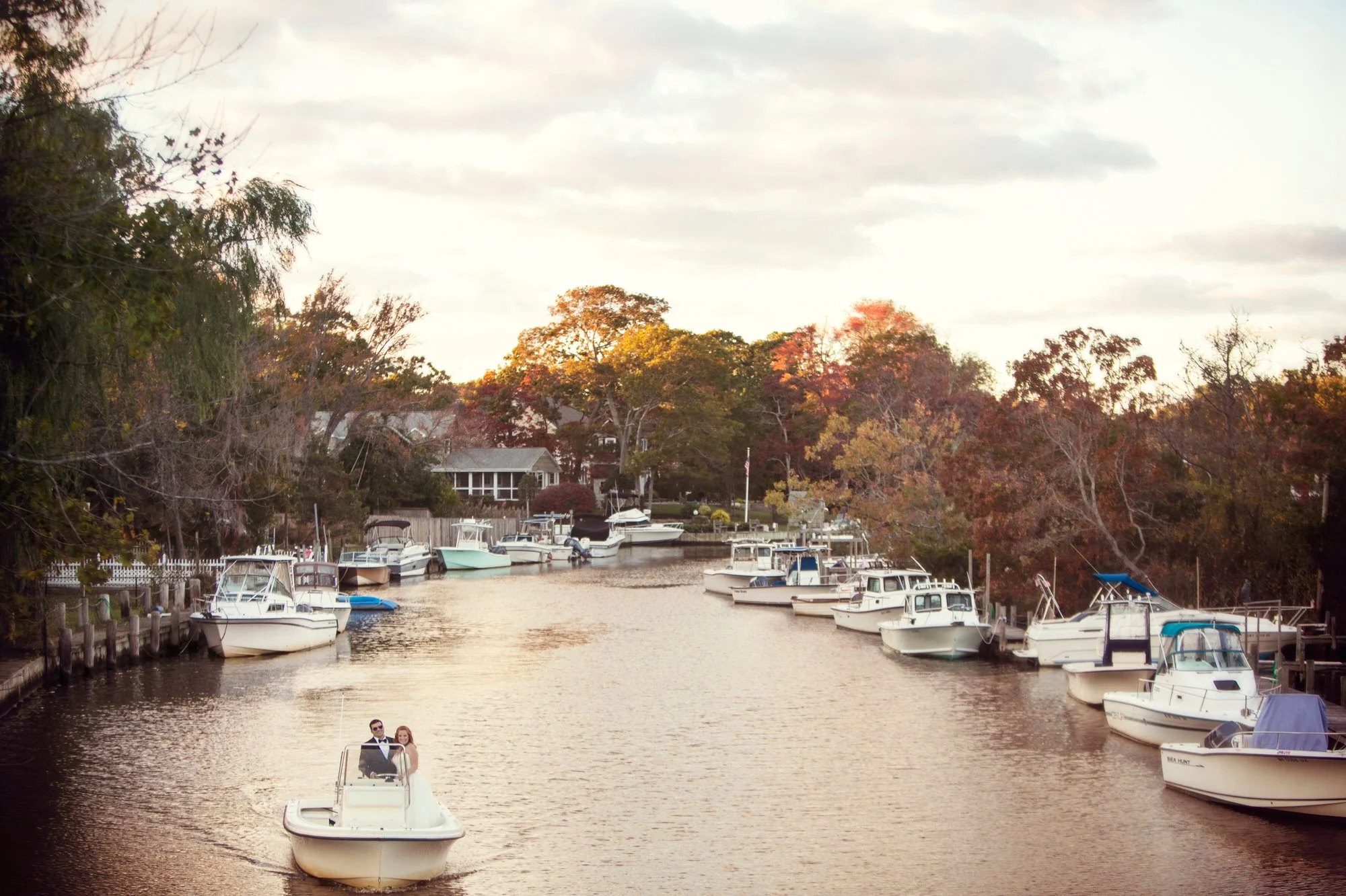 A small boat with a couple in wedding attire sailing in a canal lined with boats and trees during sunset.