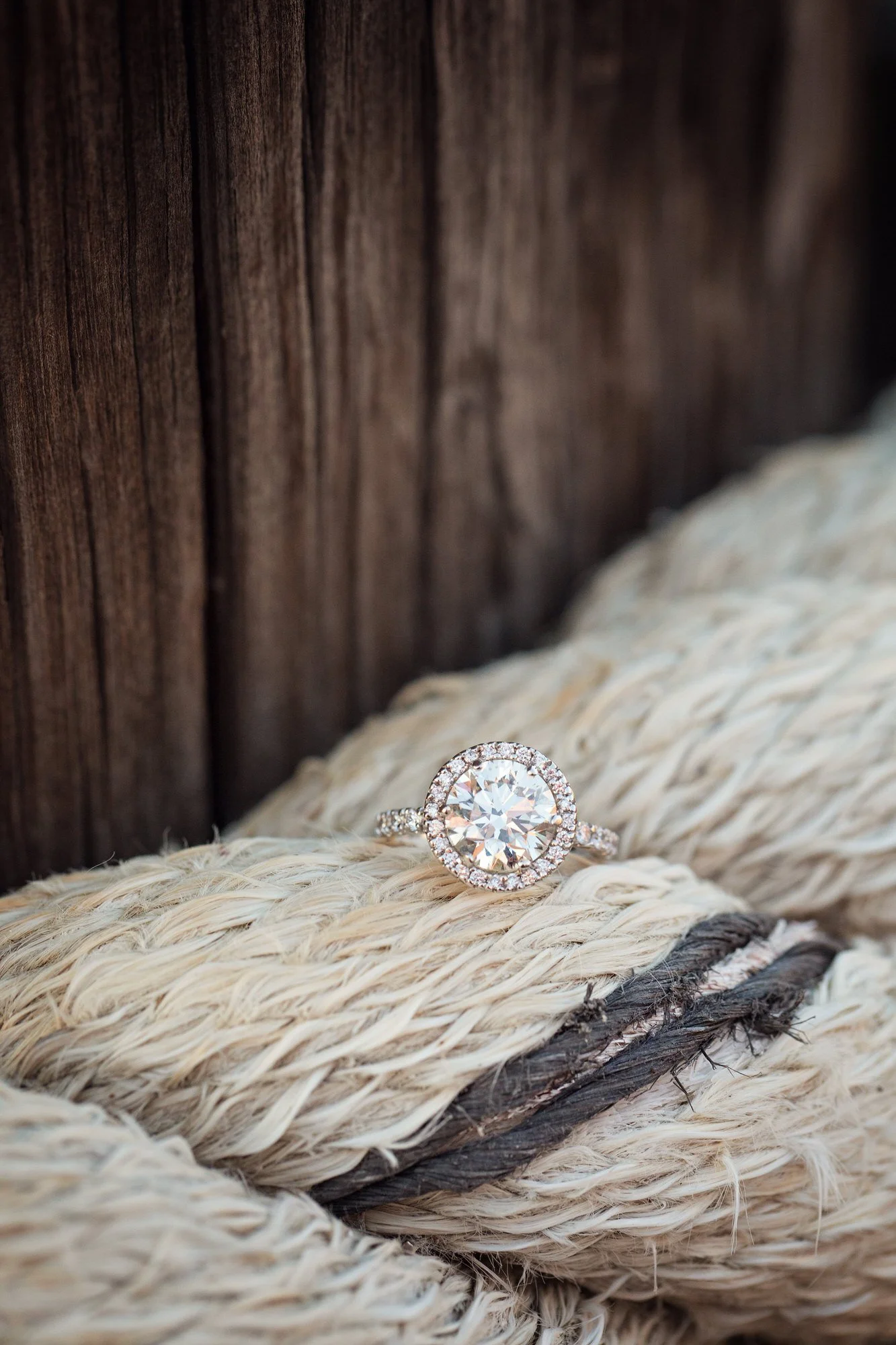 A diamond engagement ring with a halo setting rests on thick white rope against a wooden background.