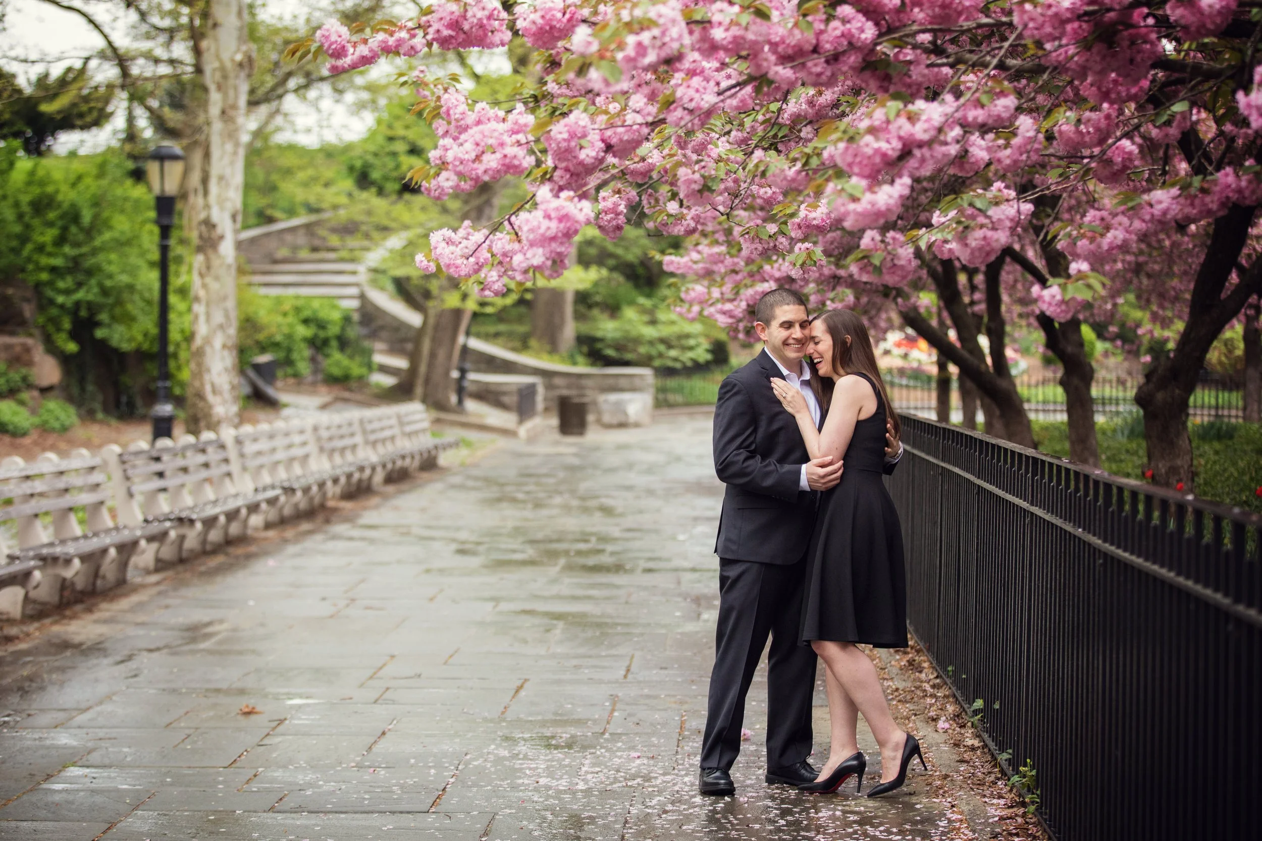 A couple in formal attire sharing a joyful moment in a park with pink flowering trees and a paved pathway.
