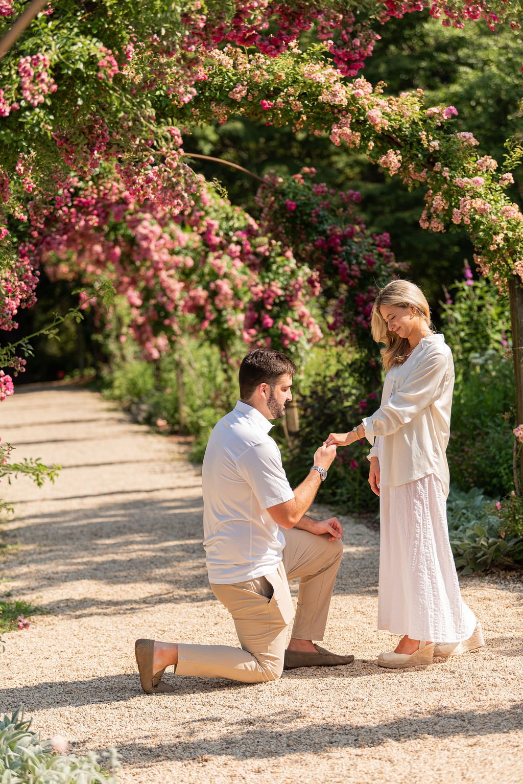 A man proposing marriage to a woman in a garden with pink and purple flowers.