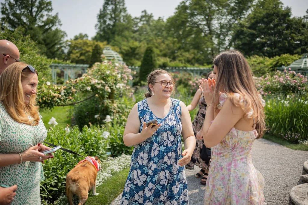 Group of women smiling and chatting in a garden, with a woman in a blue floral dress holding a phone, and another woman in a pastel dress covering her mouth in surprise, while a dog is present nearby.