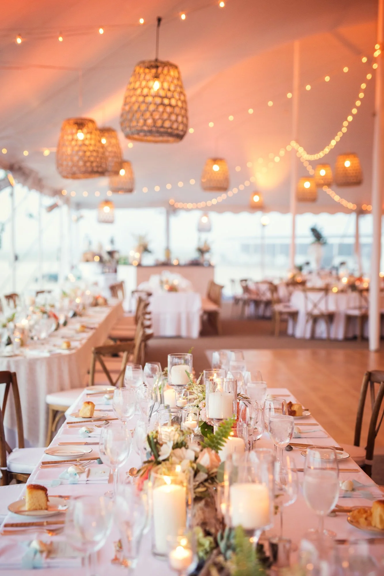 Elegant dining setup inside a decorated event tent with string lights, wicker pendant lamps, and tables adorned with candles, floral arrangements, and place settings for a wedding or celebration.