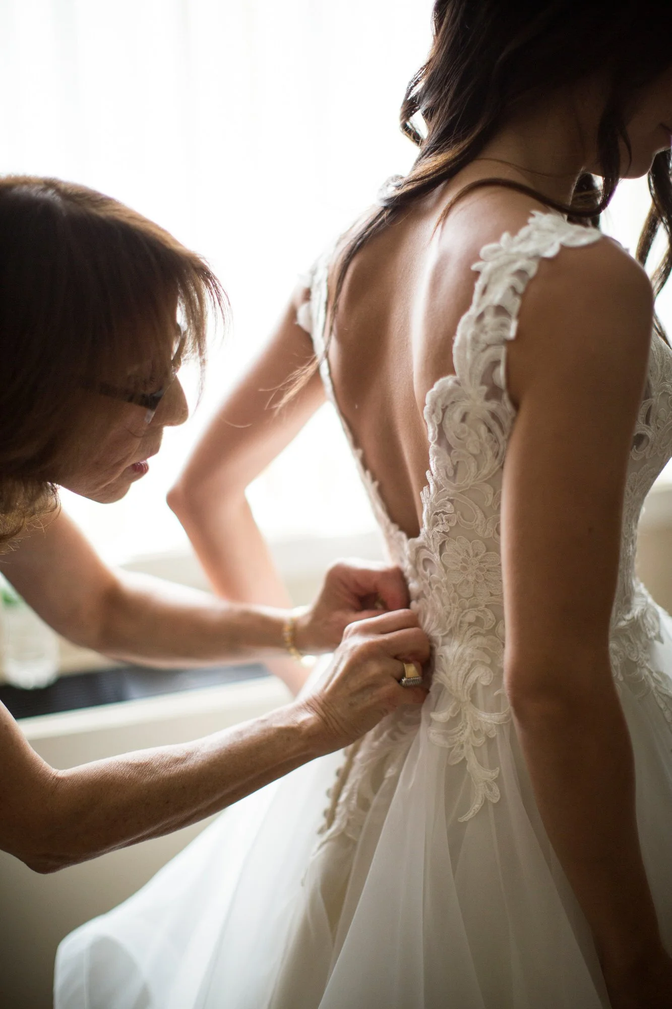 A woman in a wedding dress is being helped into her gown by another woman, possibly a family member or a stylist.