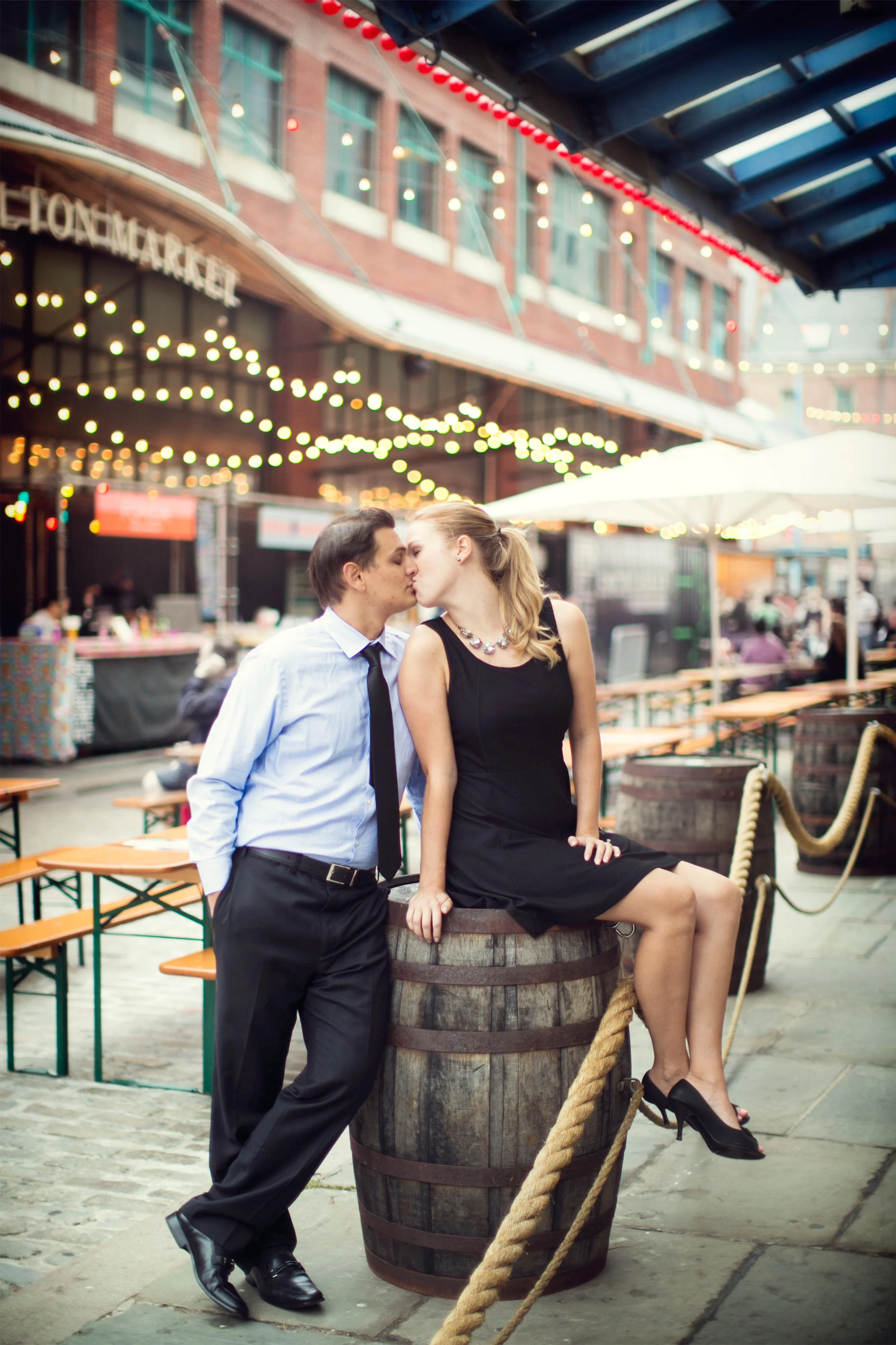 A man and woman sharing a kiss in an outdoor urban setting with string lights, restaurant tables, and a building in the background.