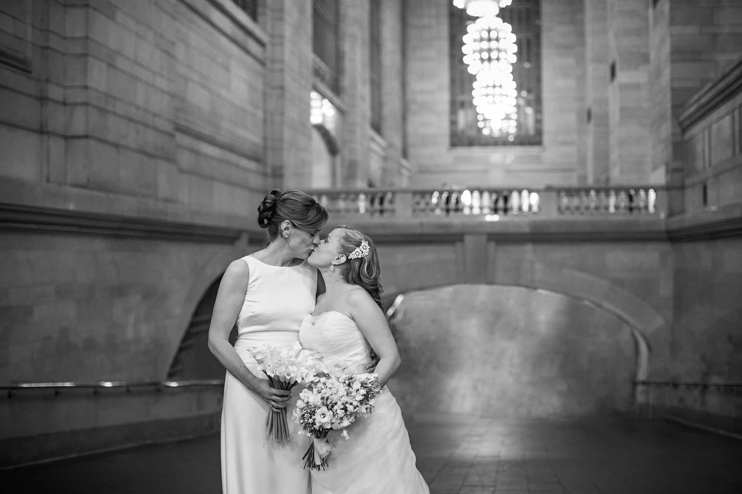 Two brides sharing a kiss in a grand, historic building with high ceilings, stone walls, and large windows; one bride with an updo hairstyle and the other with a floral headpiece, both holding bouquets.