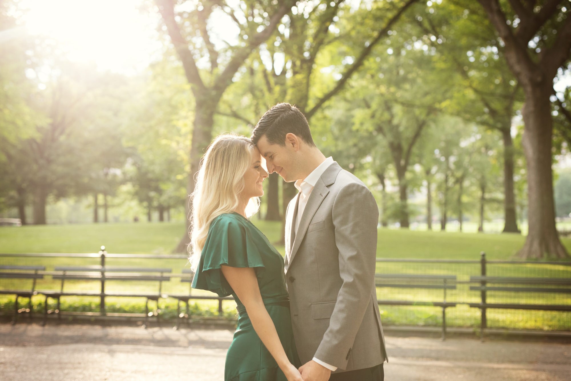 A couple in formal attire standing close in a park with tall trees and green grass, their foreheads touching and holding hands, backlit by sunlight filtering through the trees.