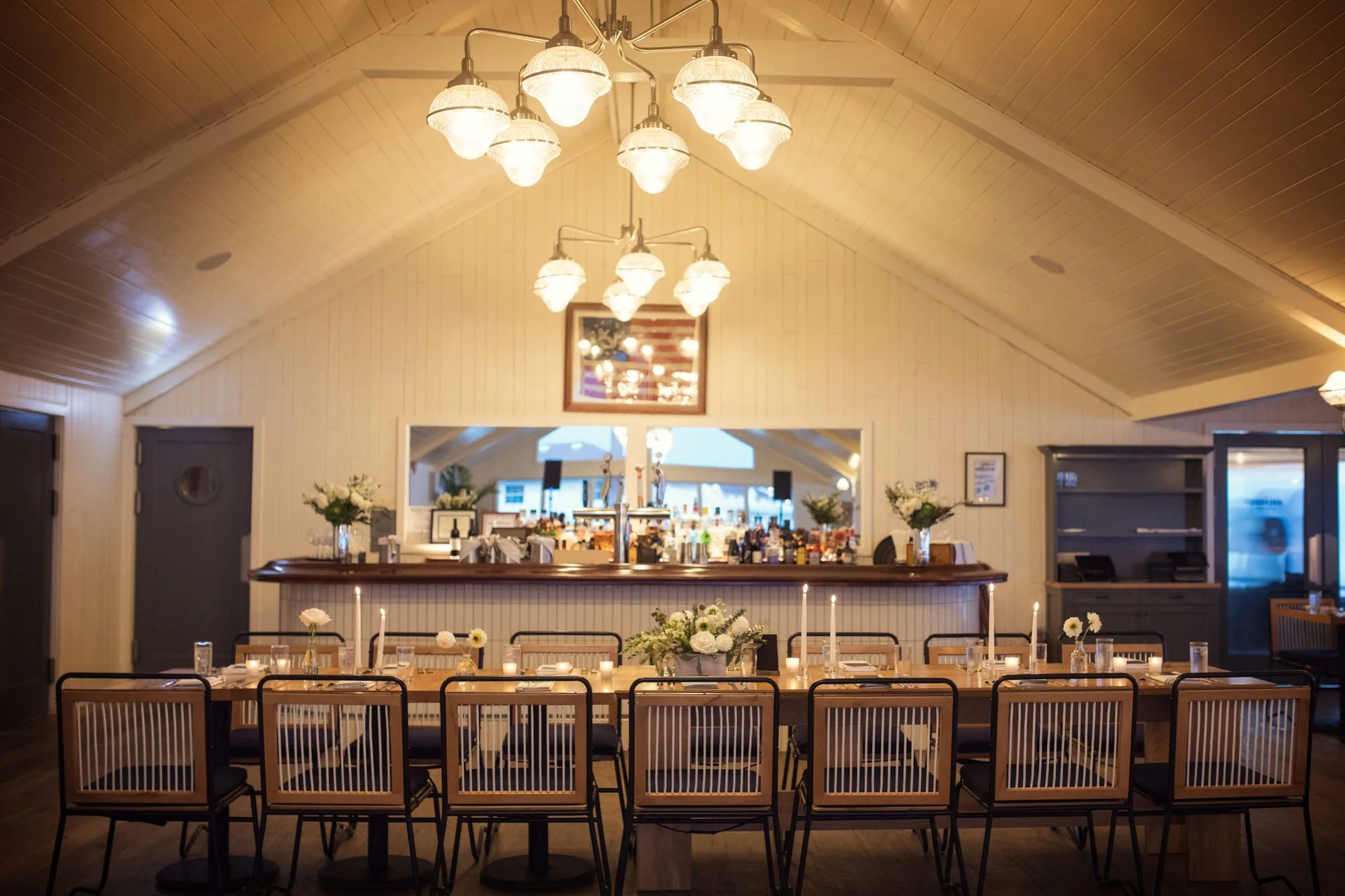 Interior of a restaurant or event space with a long dining table set with candles and flowers, a bar in the background, and chandeliers hanging from the vaulted ceiling.