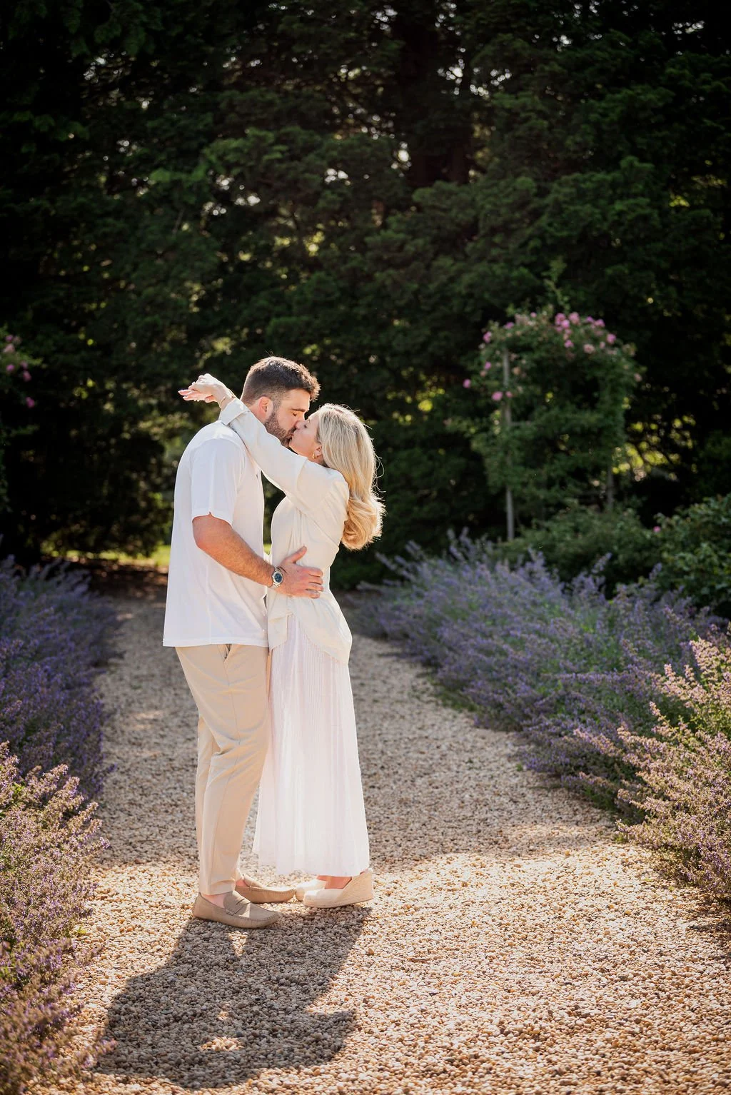 A couple kissing outdoors on a gravel path surrounded by purple flowers, with large trees and a flowering bush in the background, bathed in warm sunlight.