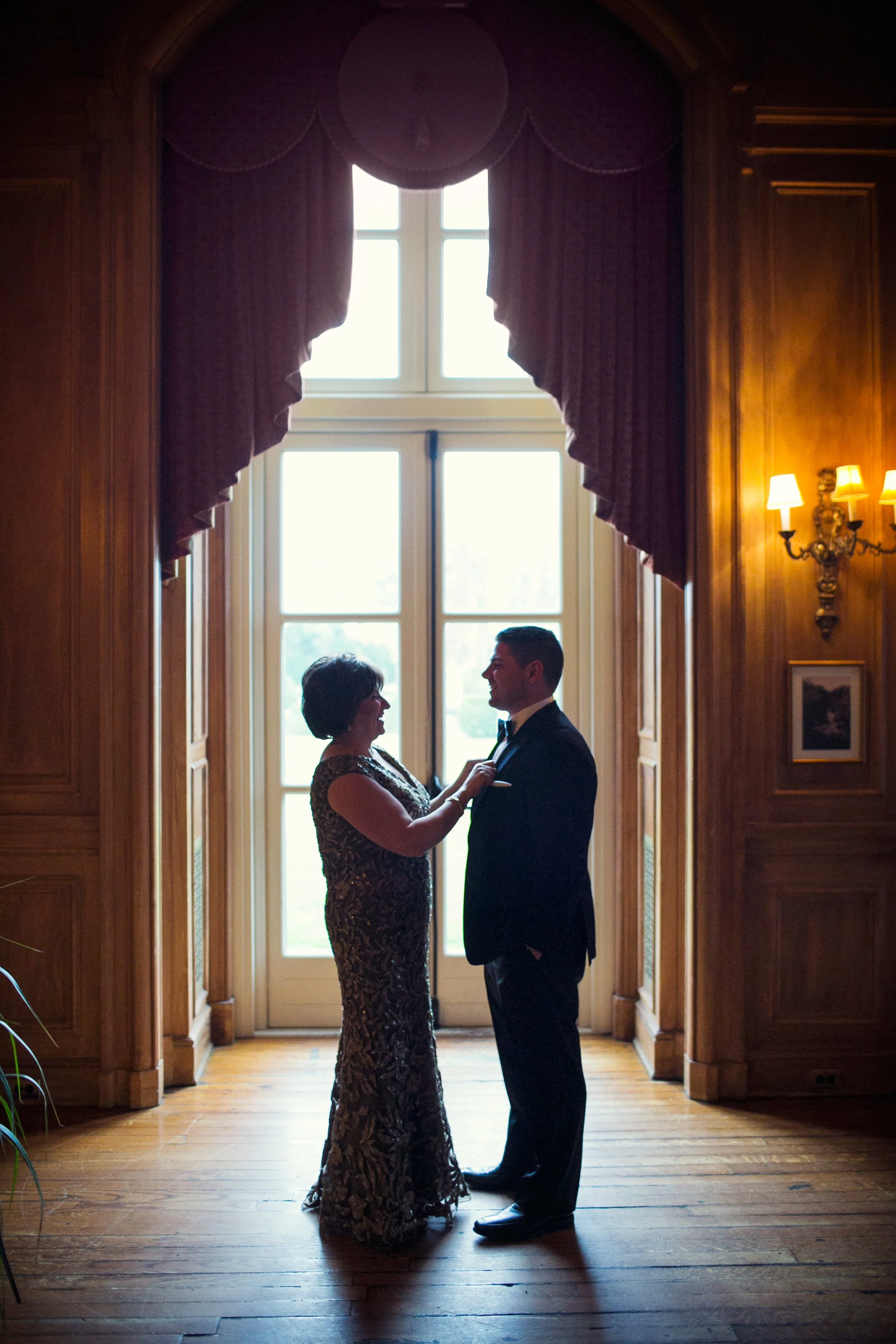 A woman in a gold and black dress and a man in a black tuxedo sharing a moment indoors near a large window with curtains.