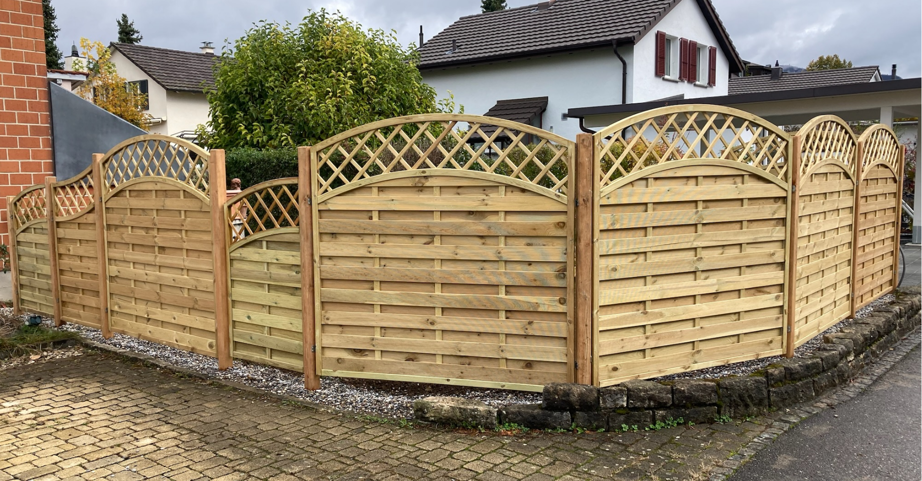 Ein umzäunter Bereich mit Holzlattenzaun vor einem Haus. Es gibt eine Pflanze und einen Gehweg mit Pflastersteinen.