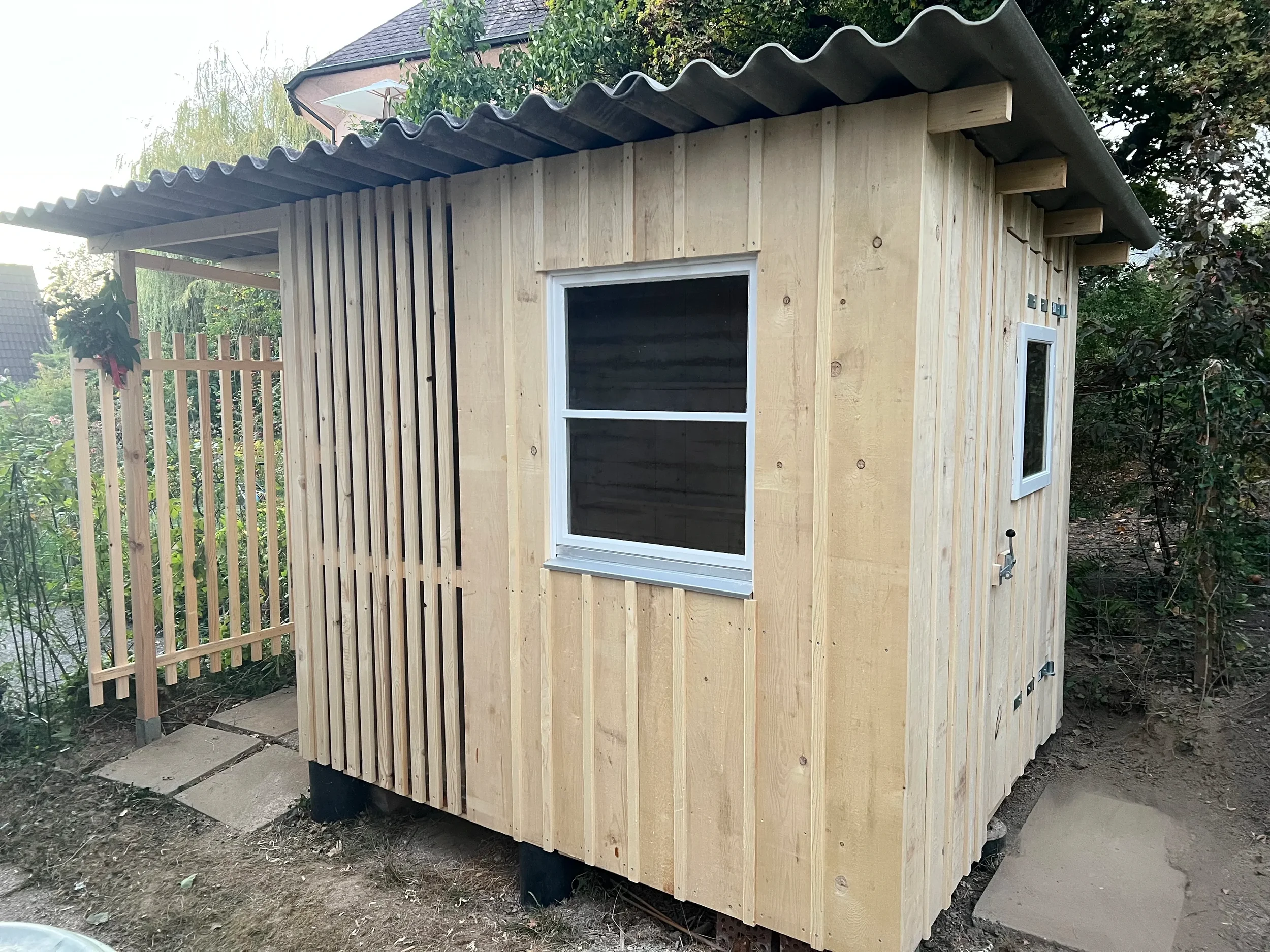 Eine kleine Holzhütte mit Fenstern und Dach, umgeben von Gartenumgebung und einer Holzlattene-Umzäunung.