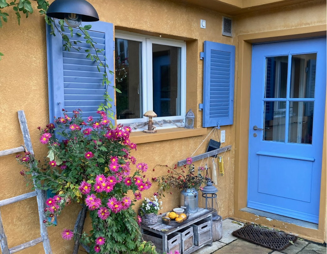 Ein gelbes Haus mit blauen Fensterläden und Tür. Blumen und Dekorationen vor dem Eingang, darunter eine große Blume, eine alte Leiter, ein kleiner Tisch mit Kerzen und Dekorationen, sowie Gartenaccessoires.