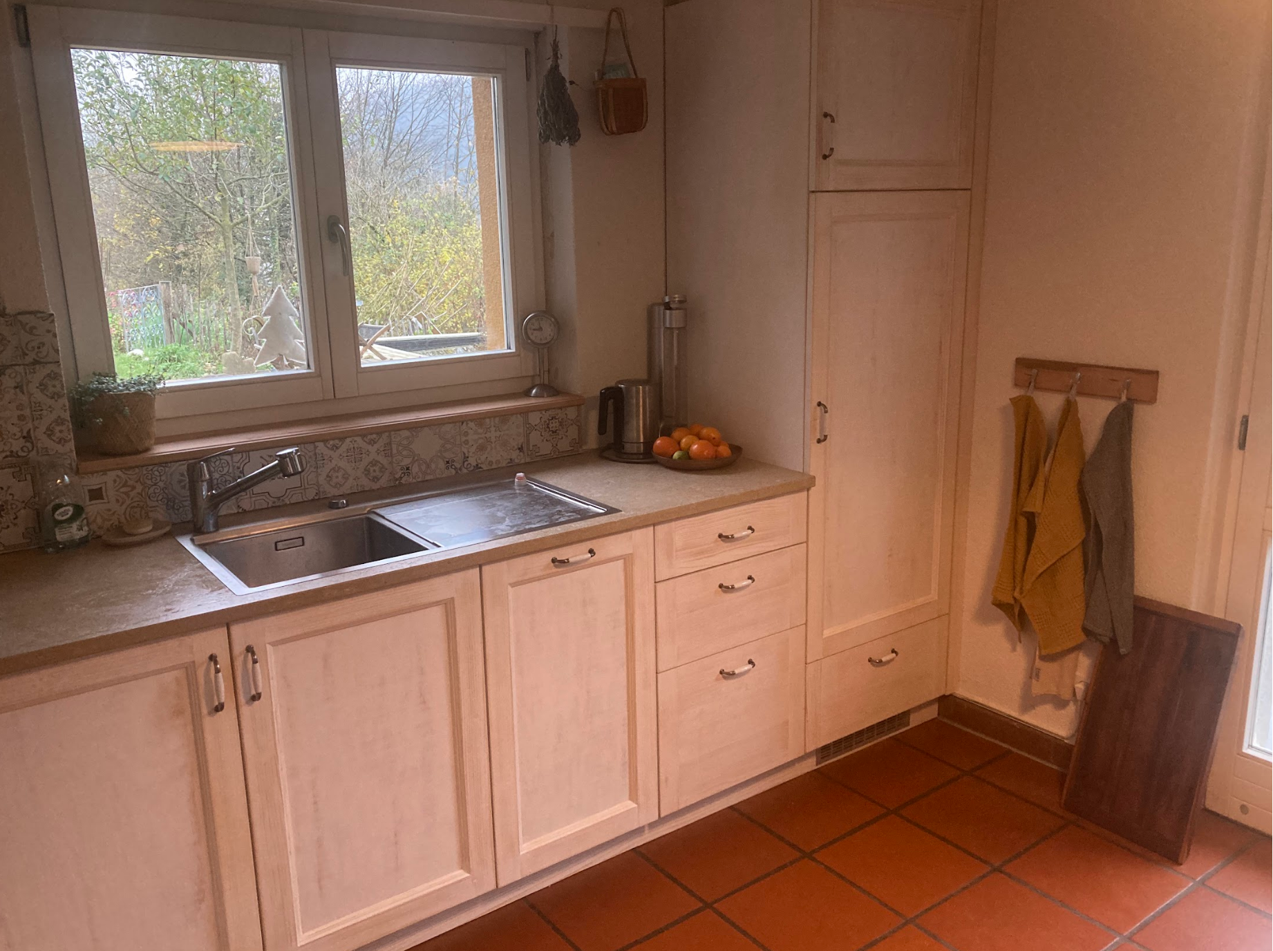 Küchenraum mit Fenster, Spüle, Wasserkocher, Obst in einer Schüssel, Handtücher an der Wand, Holzschränke und Fliesenboden.