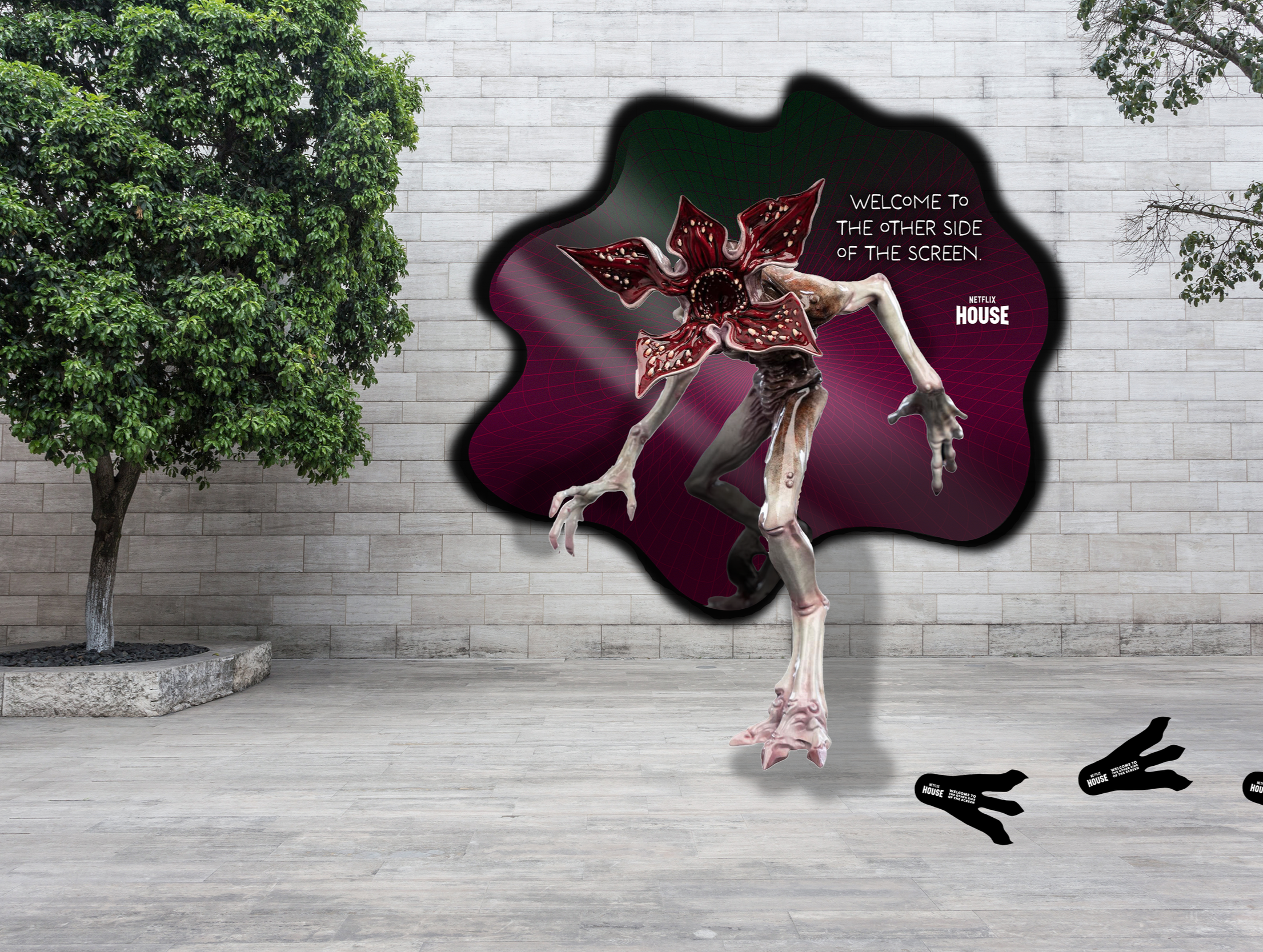 The Demogorgon emerging from a dark, cloud-shaped portal containing the text, 'Welcome to the other side of the screen.' There are several black footprints with the word 'NetflixHouse' written on them on the ground in front of the creature.