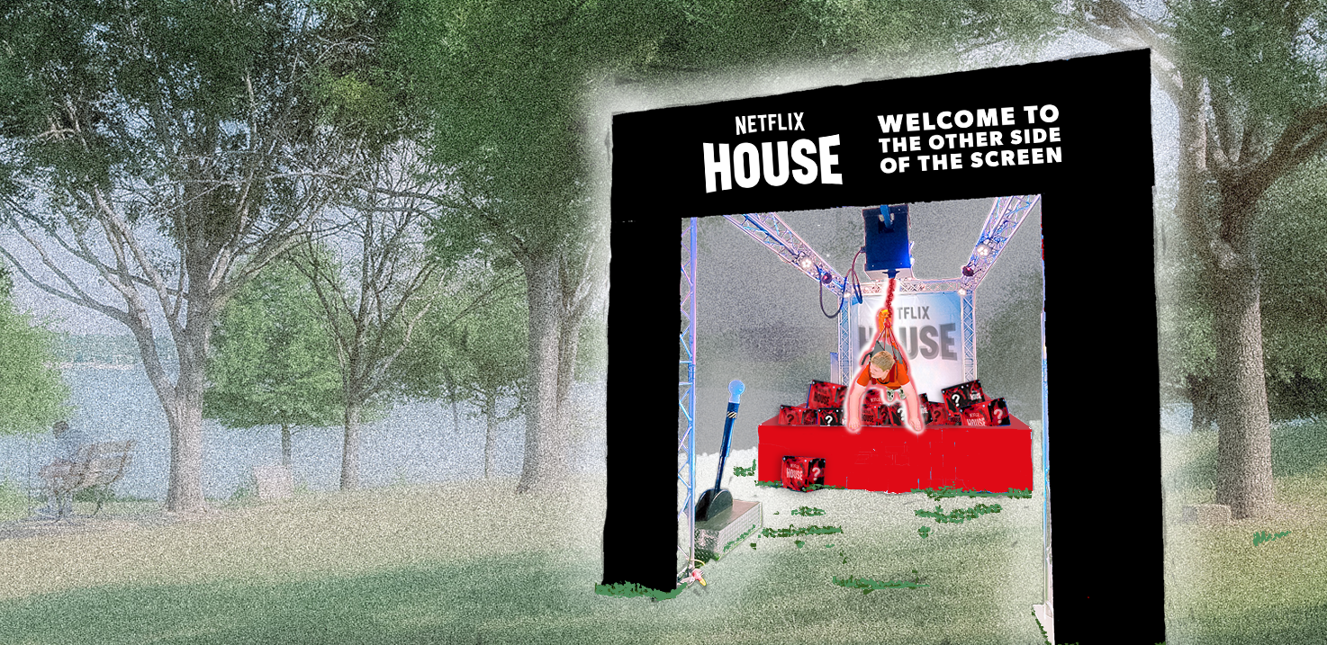 A person in a life-size prize grabber under a black rectangular sign that reads "Netflix House. Welcome to the other side of the screen" in a park with trees and grass.