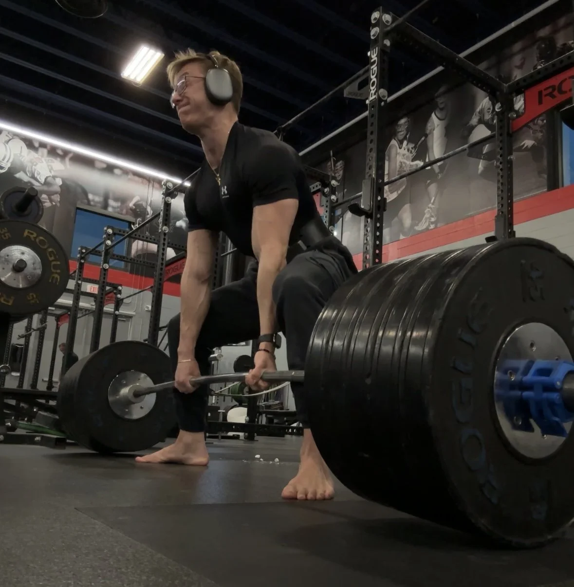 A woman with headphones and glasses performing a deadlift in a gym with black weights and a mural of athletes in the background.