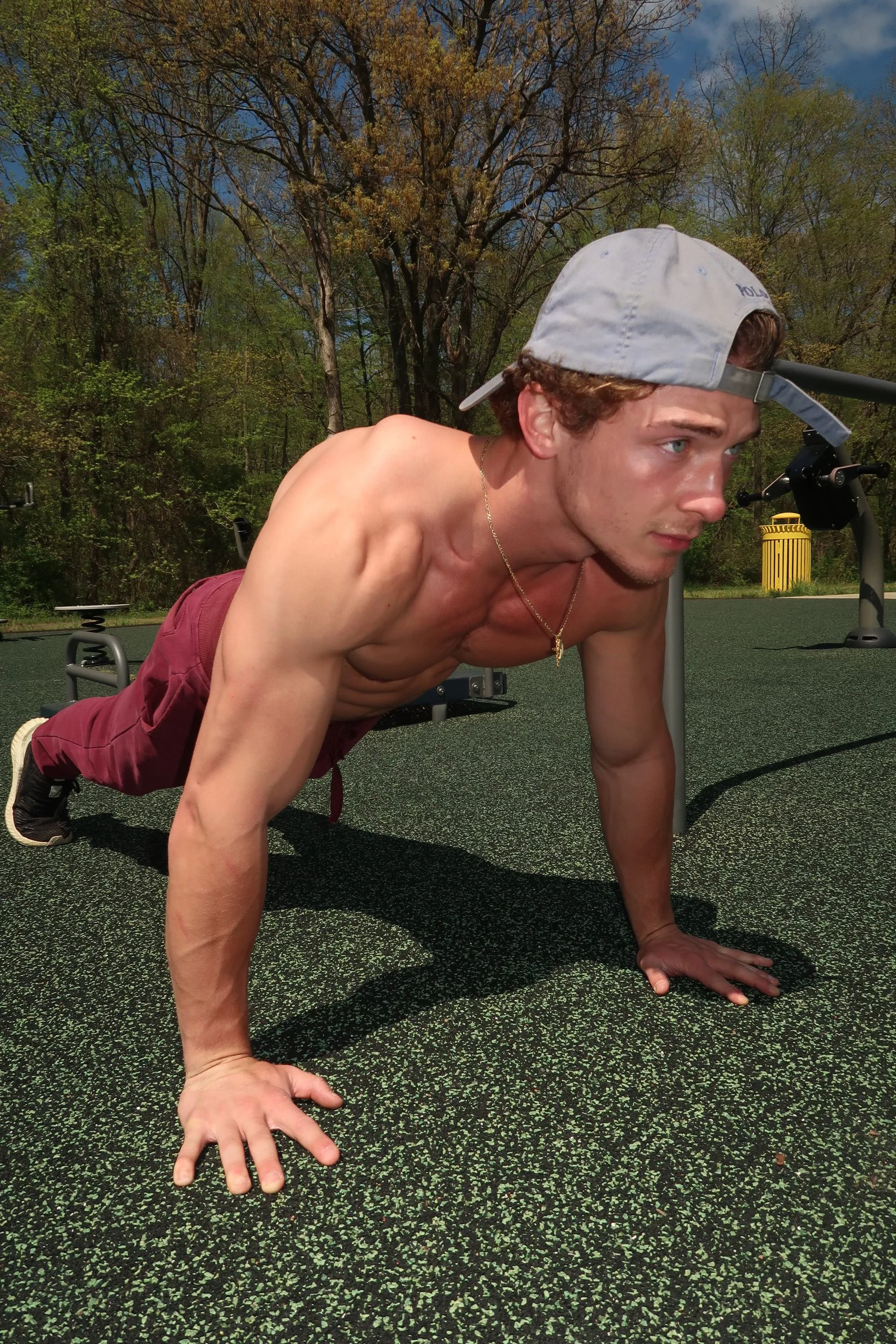 A young man doing push-ups outdoors on a rubberized surface with trees and a yellow trash bin in the background, wearing a backwards baseball cap and maroon shorts.