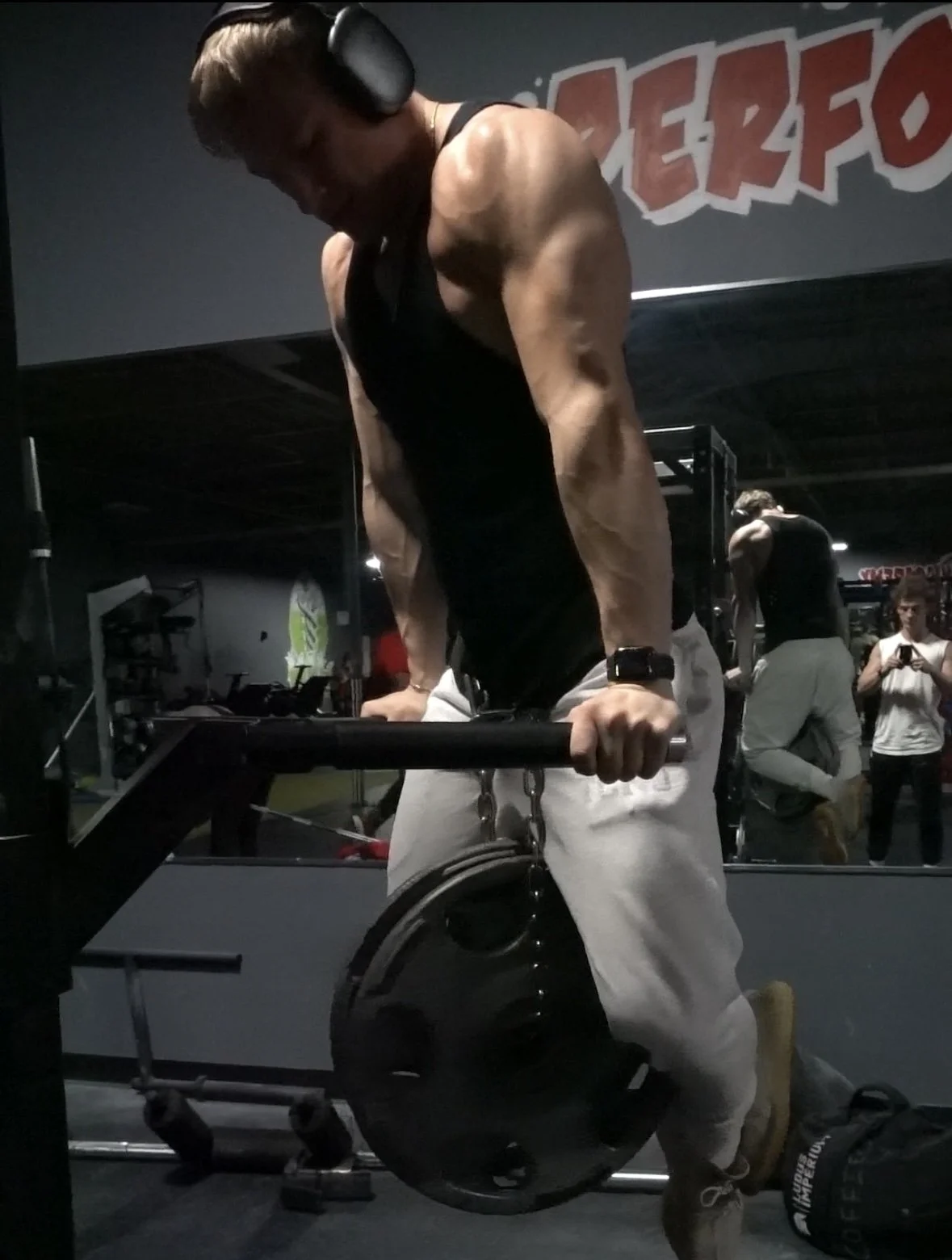 A muscular man in a black tank top and white sweatpants doing a dip exercise on parallel bars at a gym, with large weight plates attached to his waist for added resistance, and other gym members and equipment visible in the background.