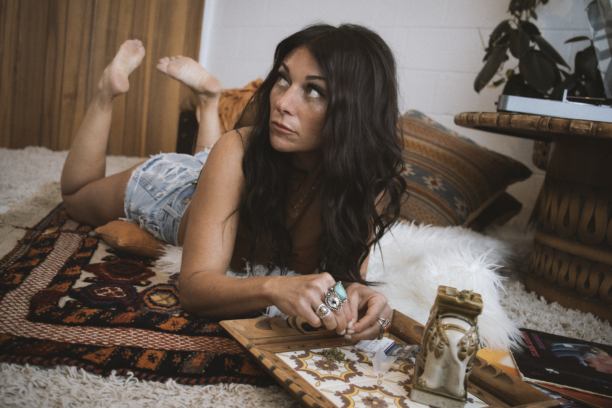 A woman with long dark hair lying on a bohemian style rug in a bedroom, propped up on her elbows, looking off to the side. She is wearing multiple rings and a necklace, with a marijuana joint and some cannabis buds on a wooden tray in front of her. T