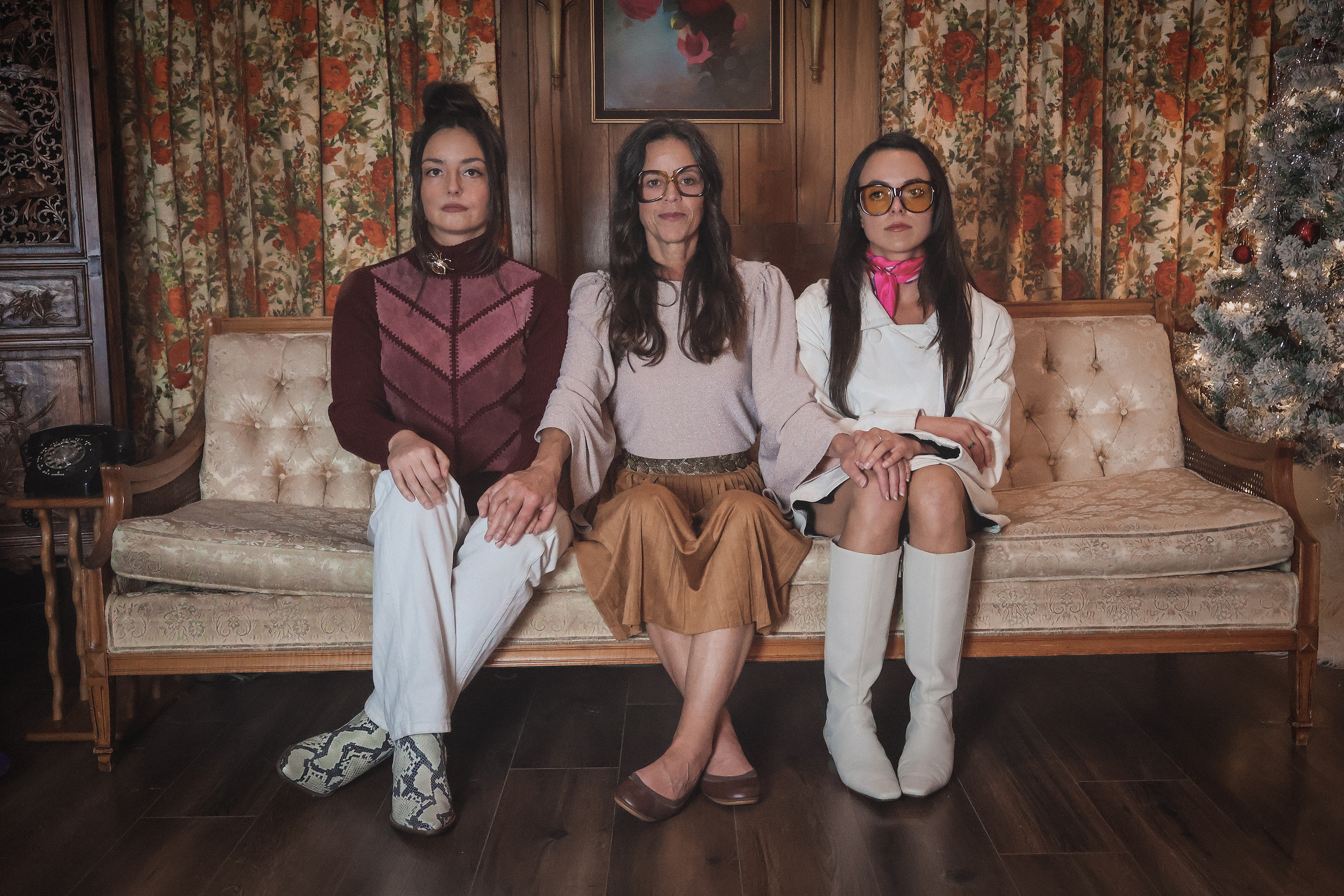 Three women sitting on a vintage sofa in a room with wood-paneled walls, floral curtains, and a Christmas tree.
